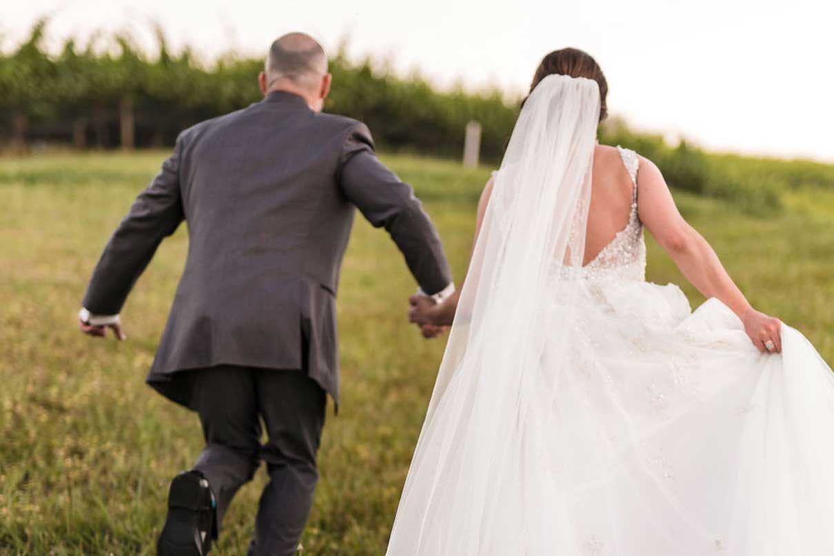 A bride and groom are running through a field holding hands.
