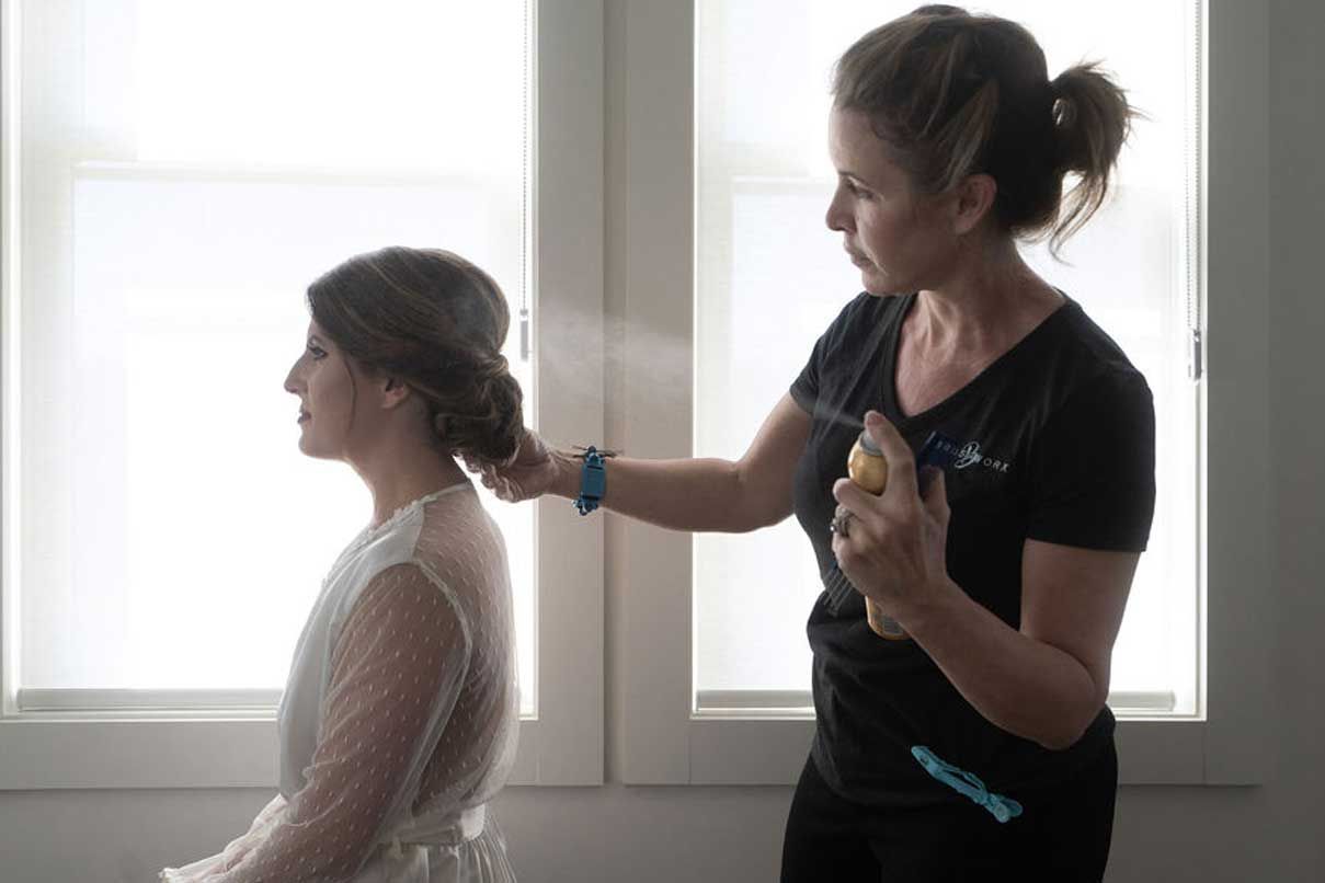 A woman is spraying a woman 's hair in front of a window.