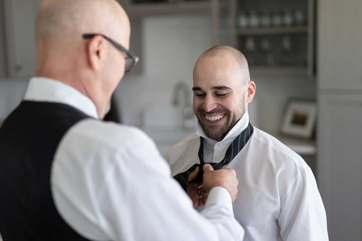 A man is helping another man tie his tie in a kitchen.