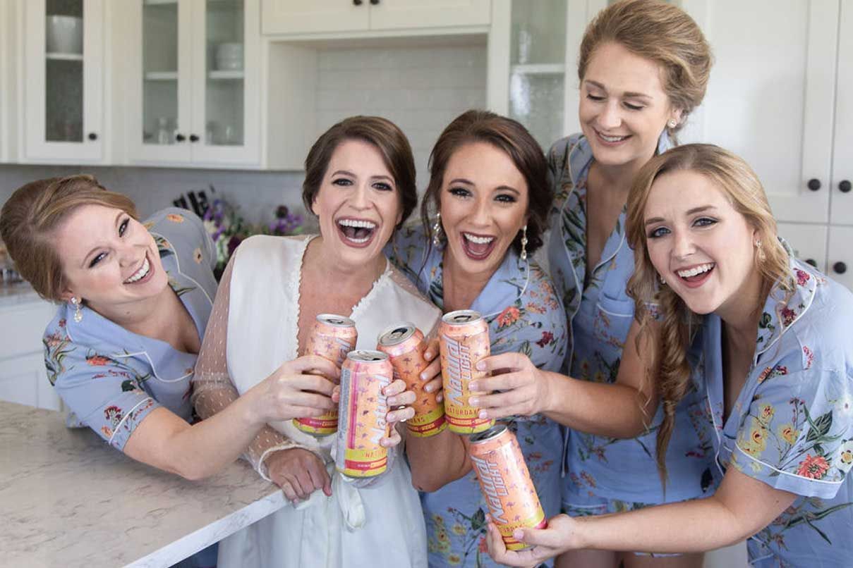 A bride and her bridesmaids are toasting with cans of soda in a kitchen.