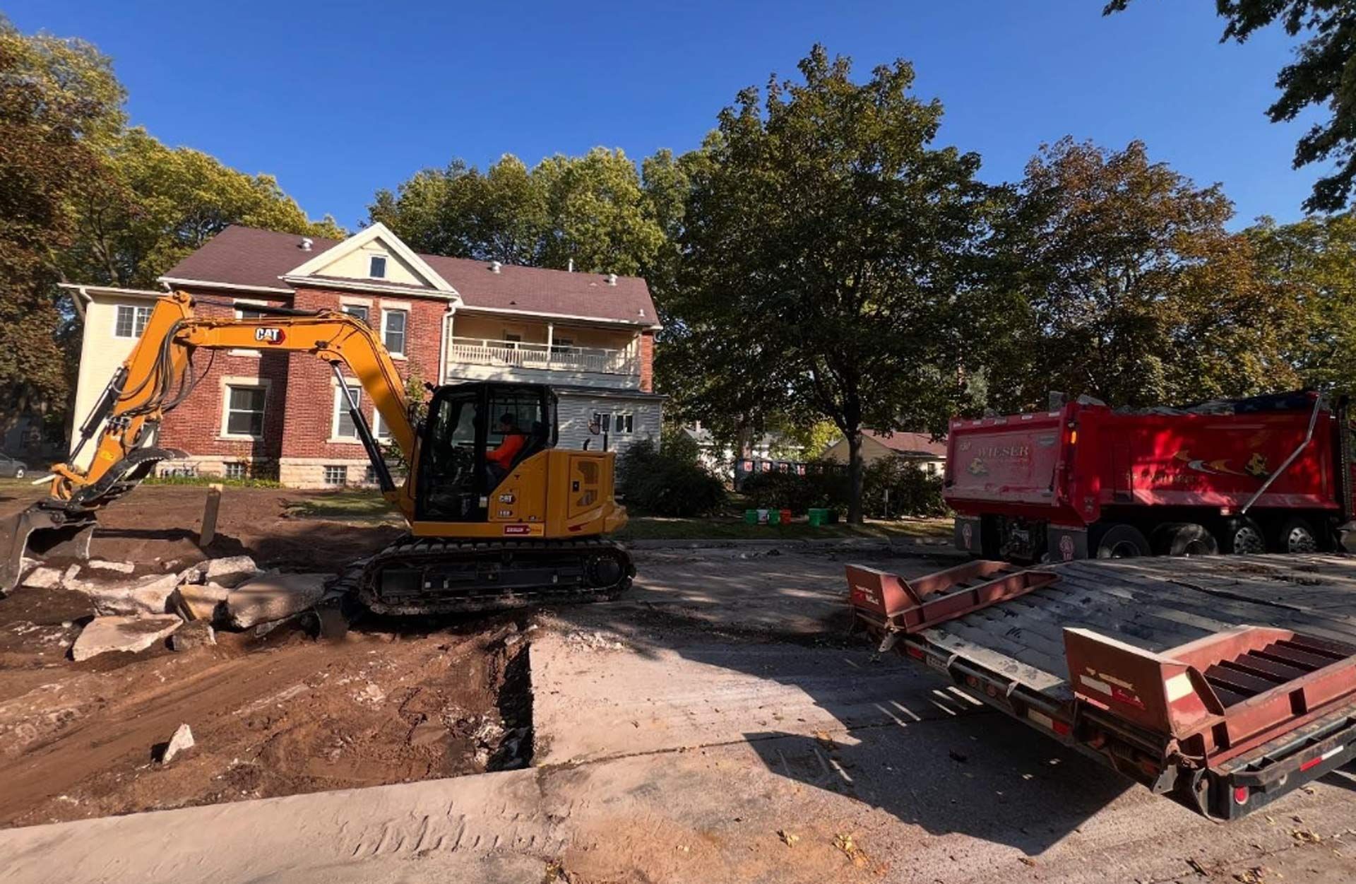 A yellow excavator is digging a hole in front of a brick house