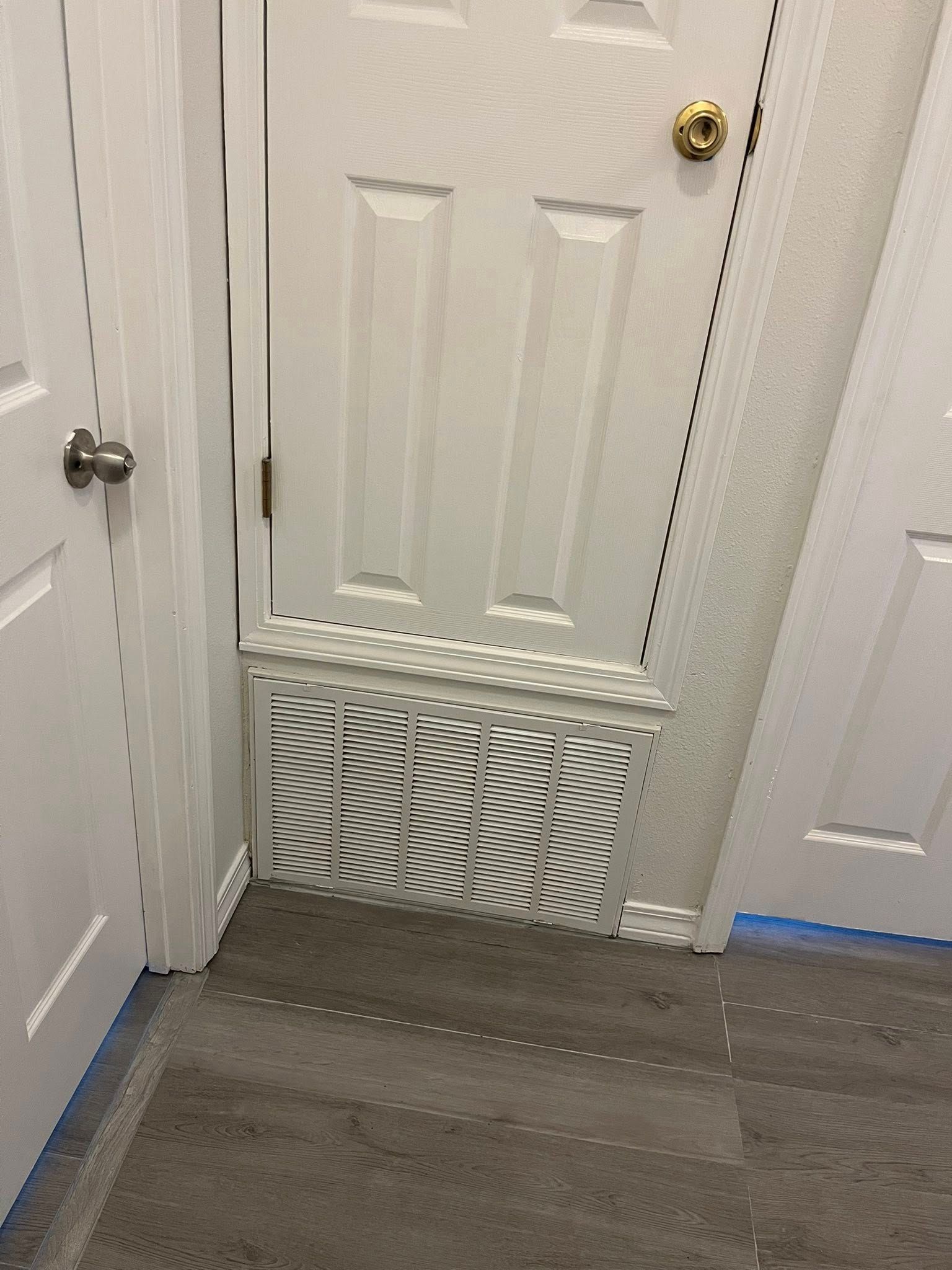 White door with decorative air vent at the bottom, gold knob, and gray flooring.