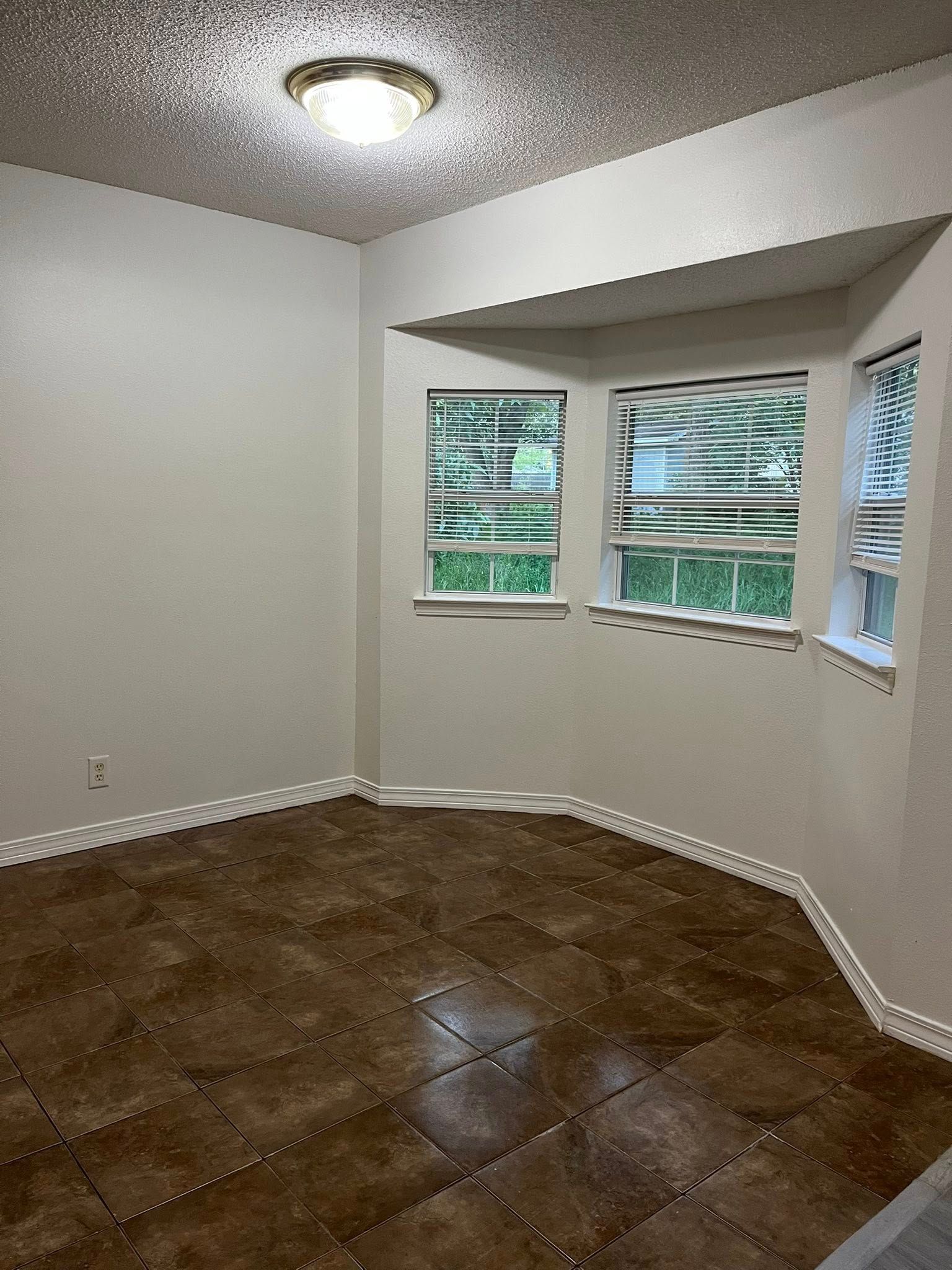 Empty room with brown tiled floor, white walls, and three windows. A ceiling light illuminates the textured ceiling.