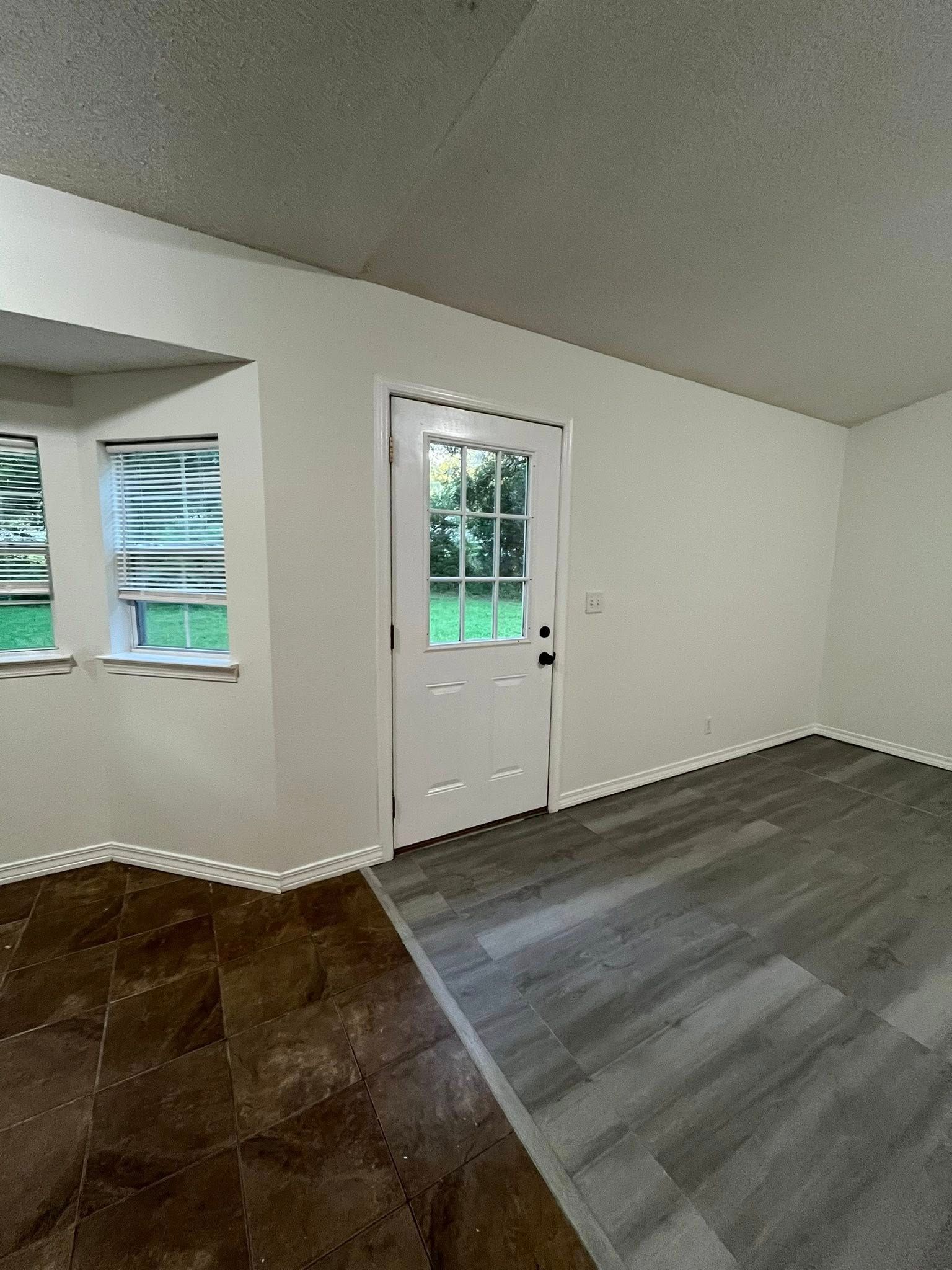 Interior view of a room with a white door, windows, and two different types of flooring: brown tile and gray wood.