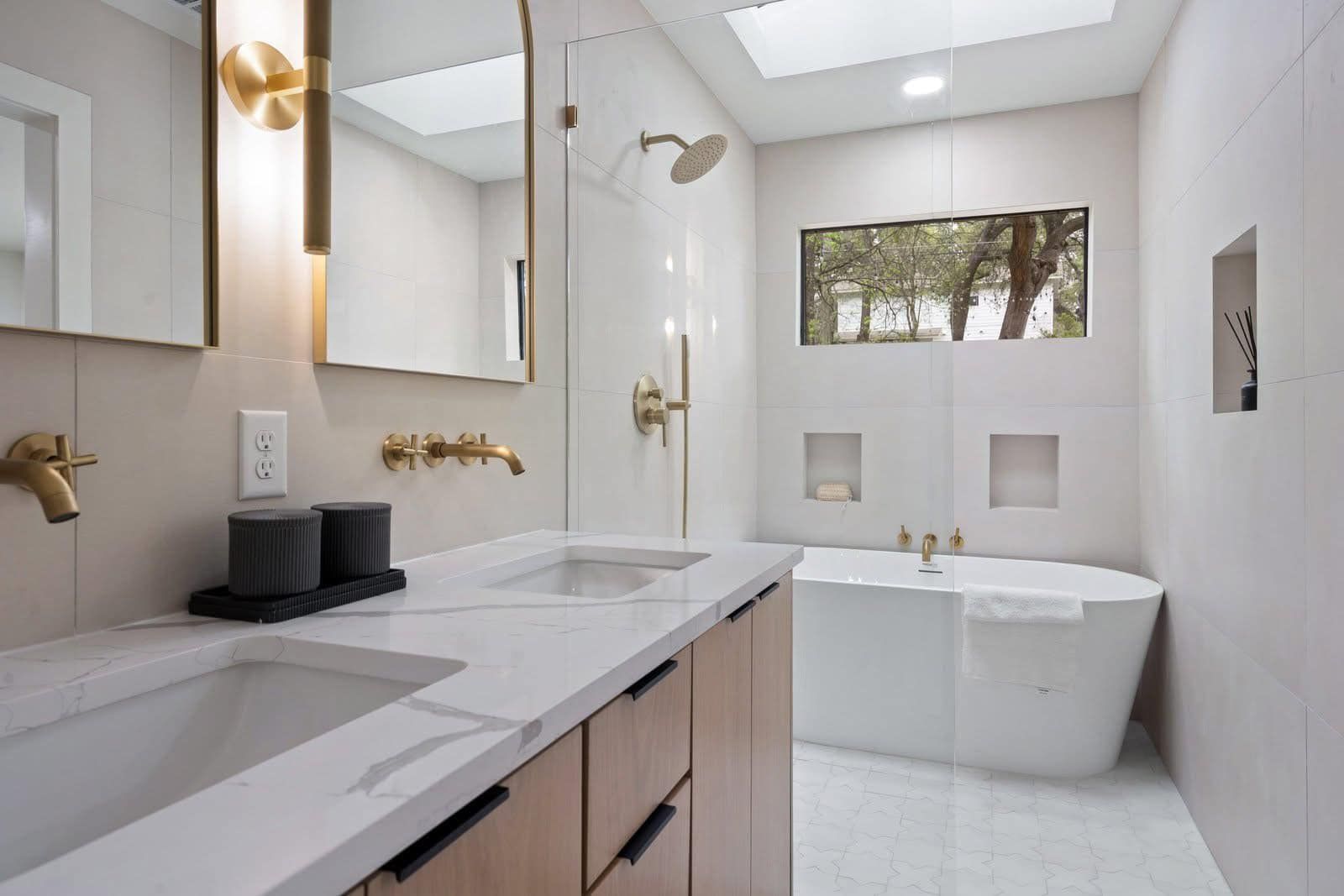 Modern bathroom with double vanity, bathtub, and natural light. Gold fixtures and white tile.