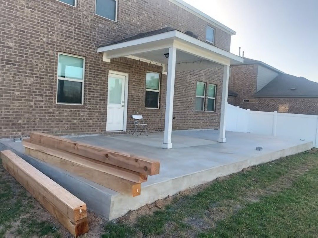 Exterior view of a brick house with a concrete patio and a white covered porch. Lumber sits in front.