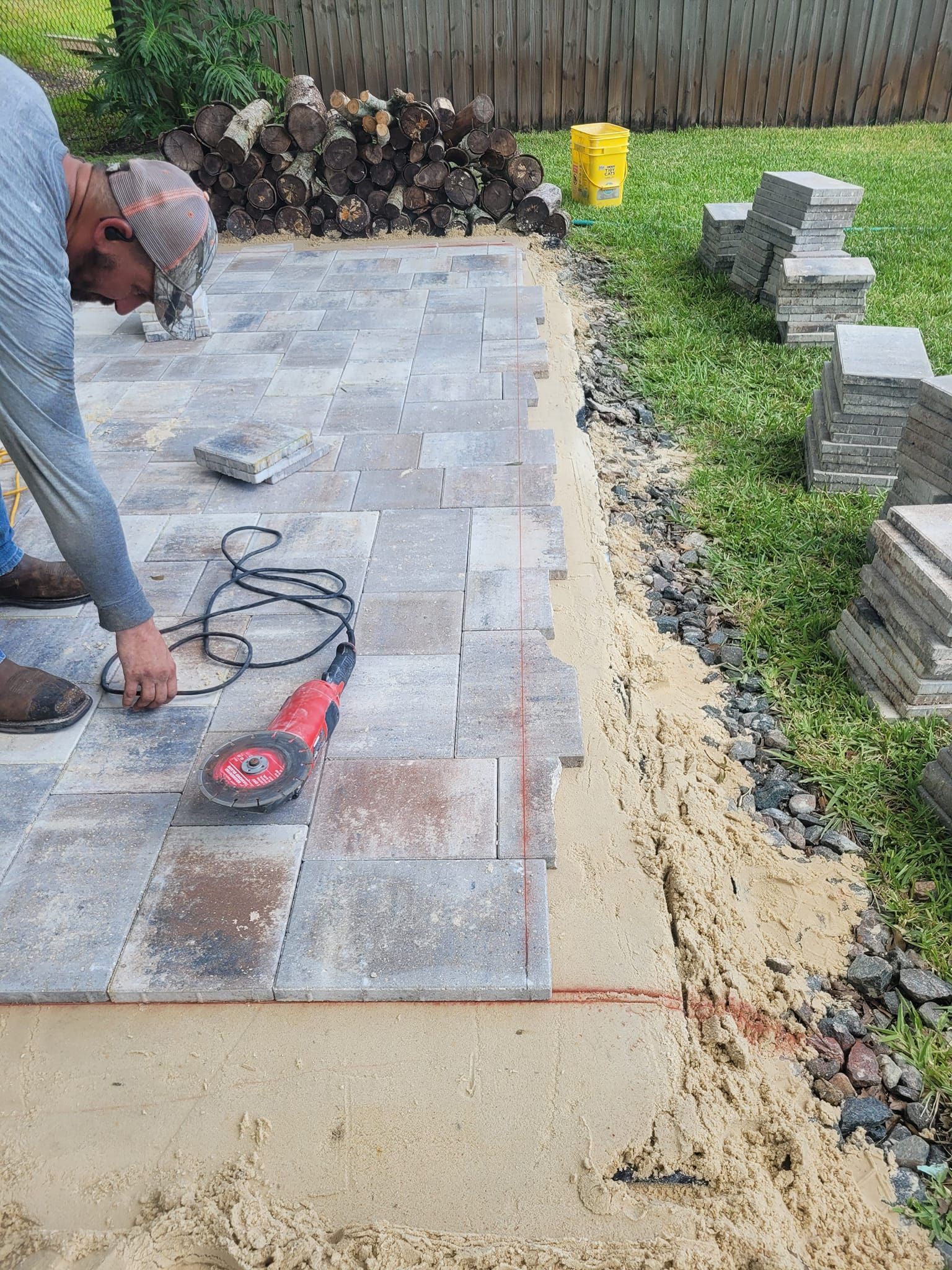Man using a power saw to cut pavers for a patio. Sand base and stone pavers are visible. Outdoors.
