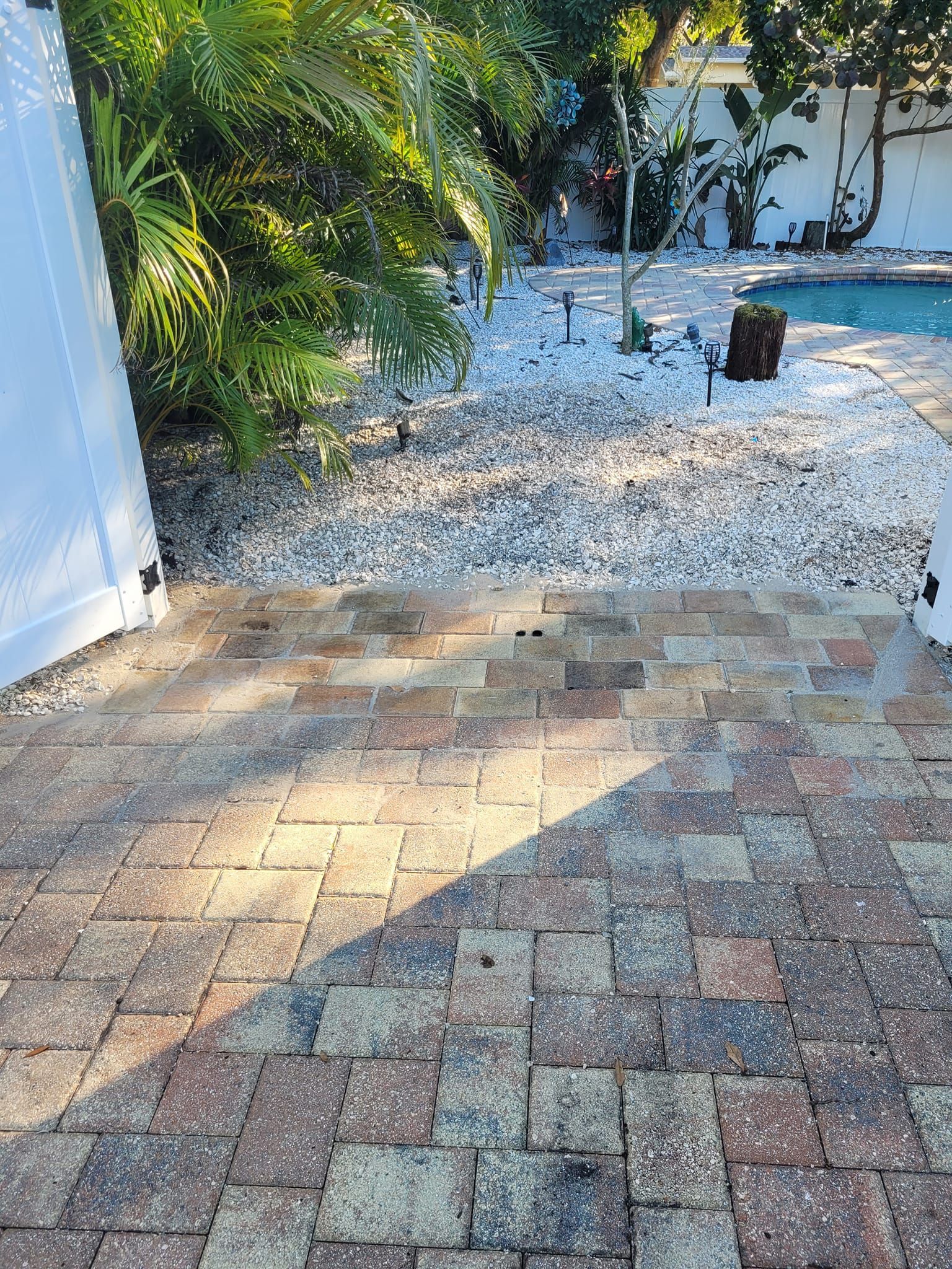 Brick patio transitioning to gravel path, leading to a pool area with trees.
