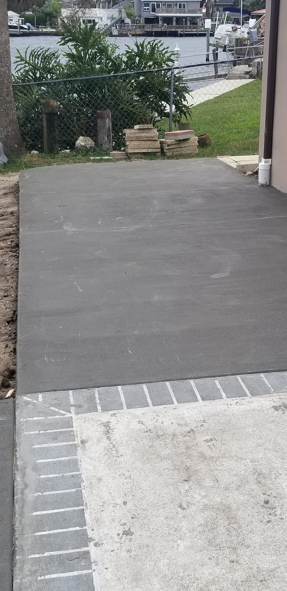Gray concrete patio with a border of gray bricks next to a gravel area, greenery, and buildings in the background.
