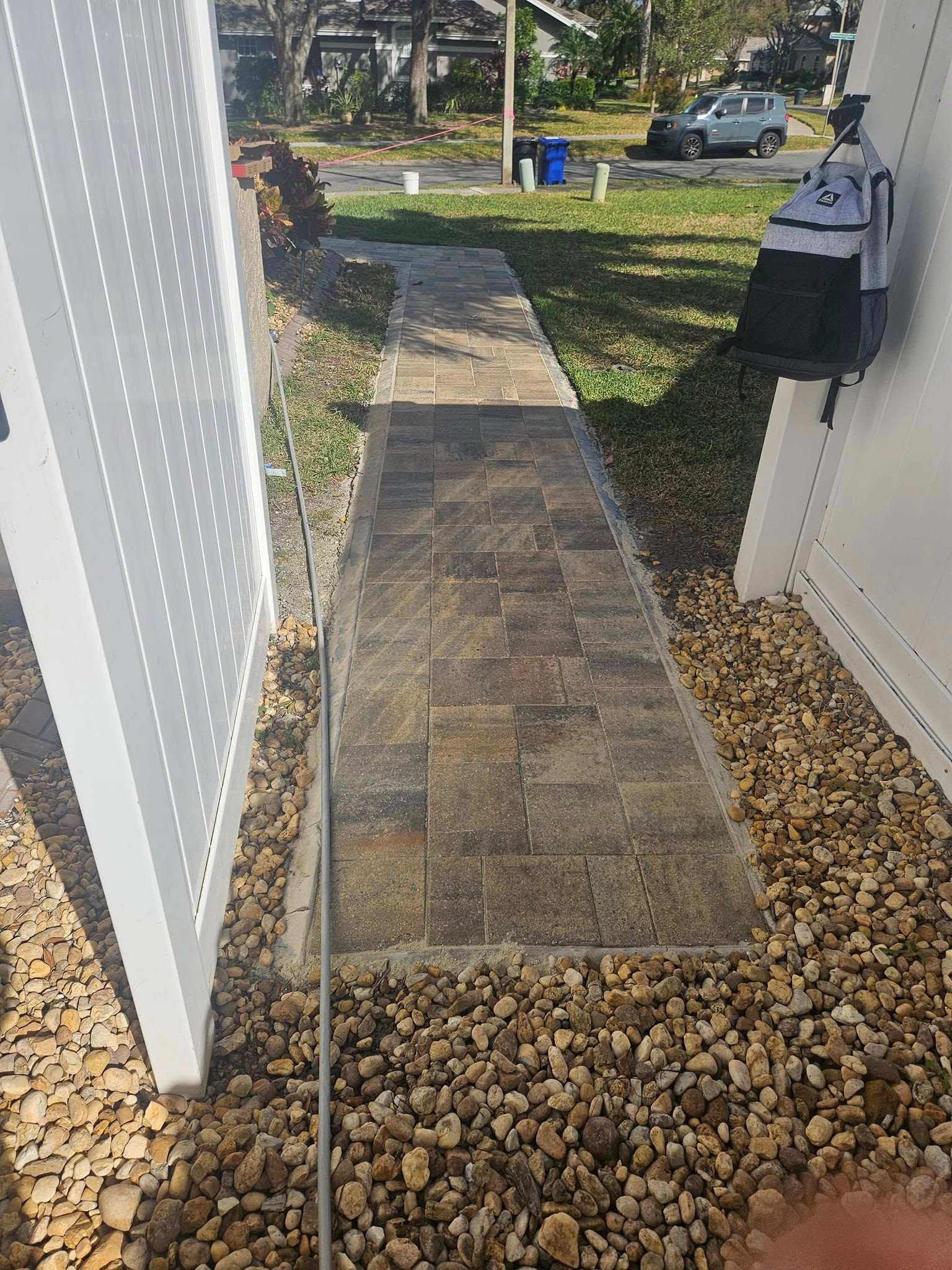 Brick walkway lined with rocks, bordered by white fence and side of a house.