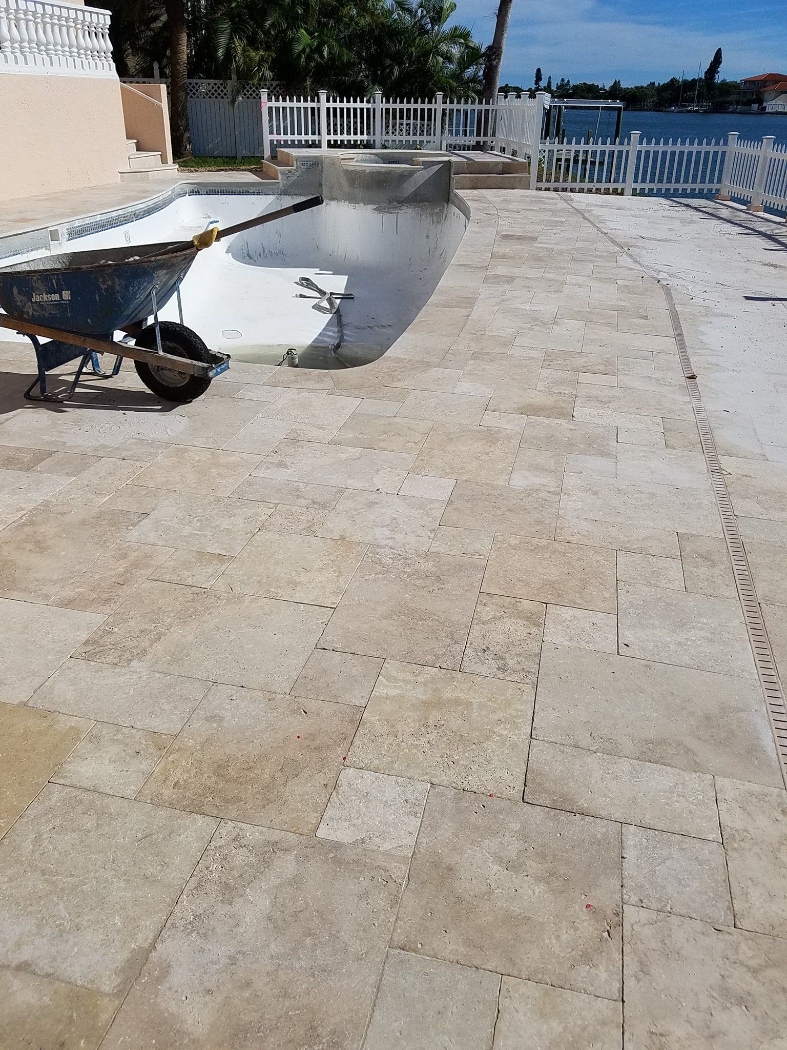 Empty swimming pool and surrounding stone patio, wheelbarrow. White fence, sunny day.