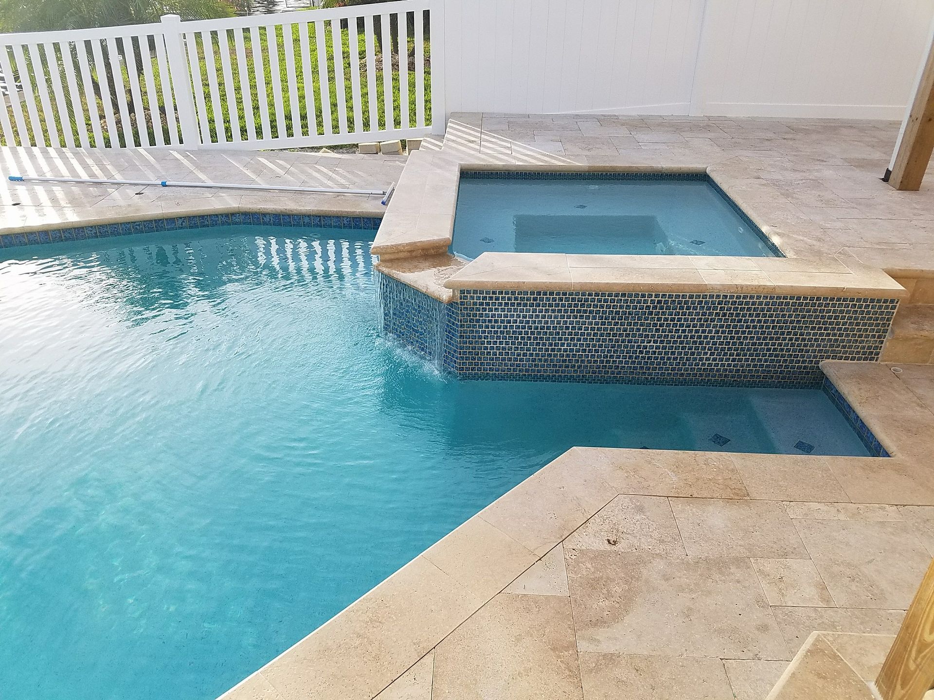 Swimming pool with attached spa, turquoise water, tan stone deck, blue tile accent, white fence.