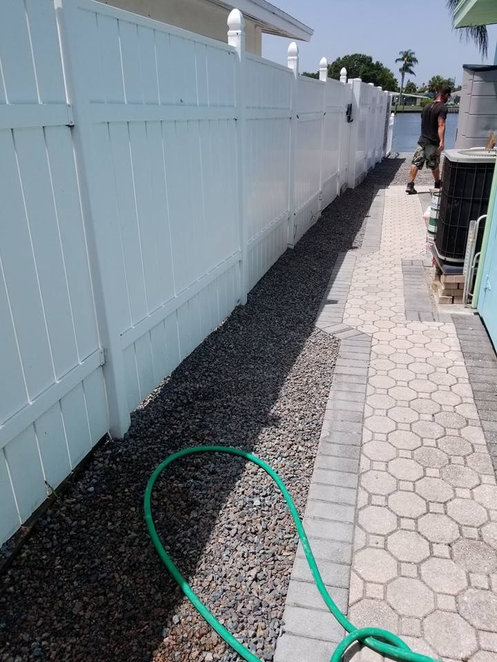 White fence next to gravel path and decorative stone walkway; a green hose lies on the ground.