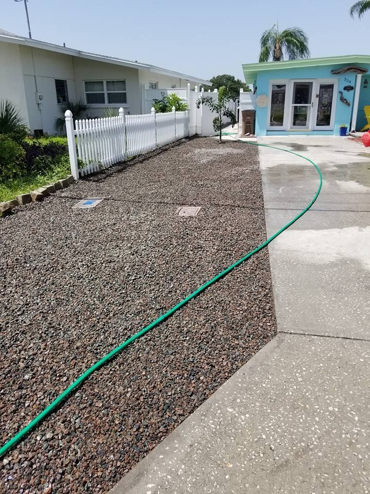 A gravel-covered yard alongside a concrete driveway with a white picket fence, a house, and a green garden hose.