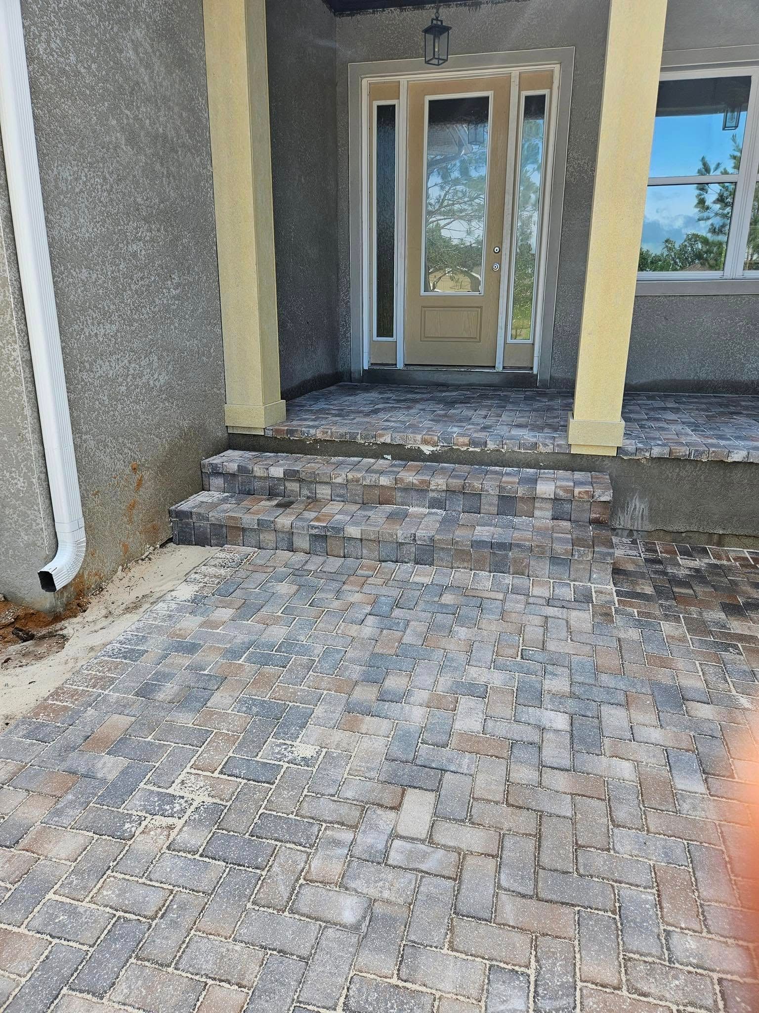 Brick paver walkway and steps leading to a house entrance with glass door. Beige pillars and gray stucco.