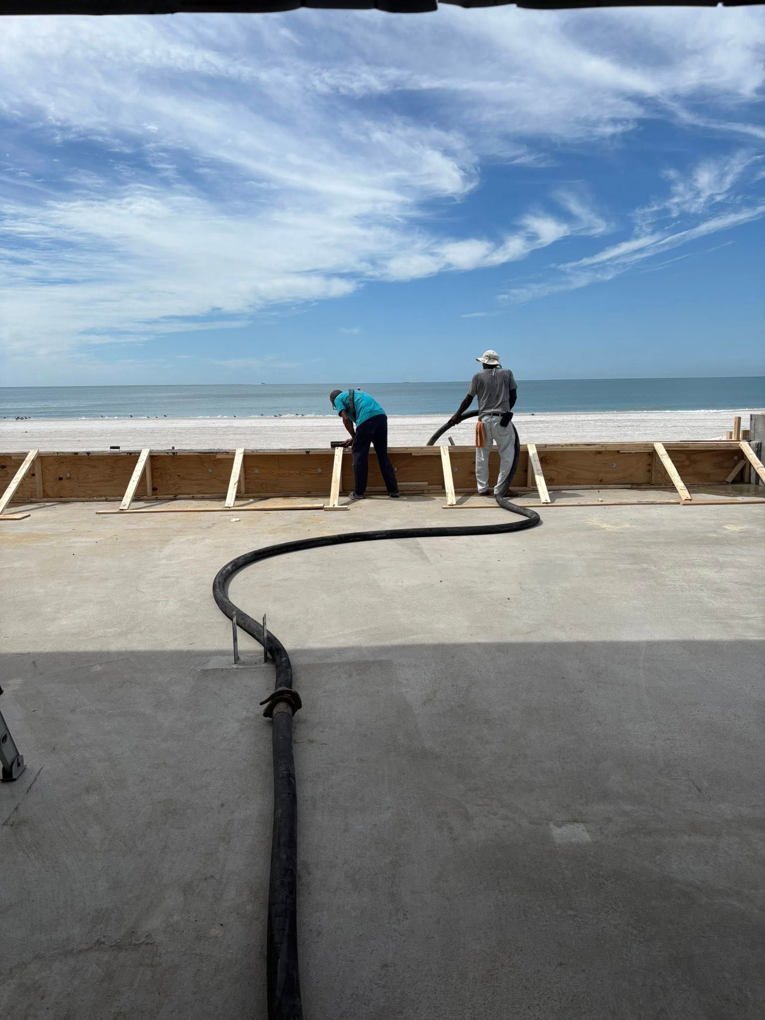 Workers pouring concrete on a beach, blue sky. A hose, wooden frames, and sea are also visible.