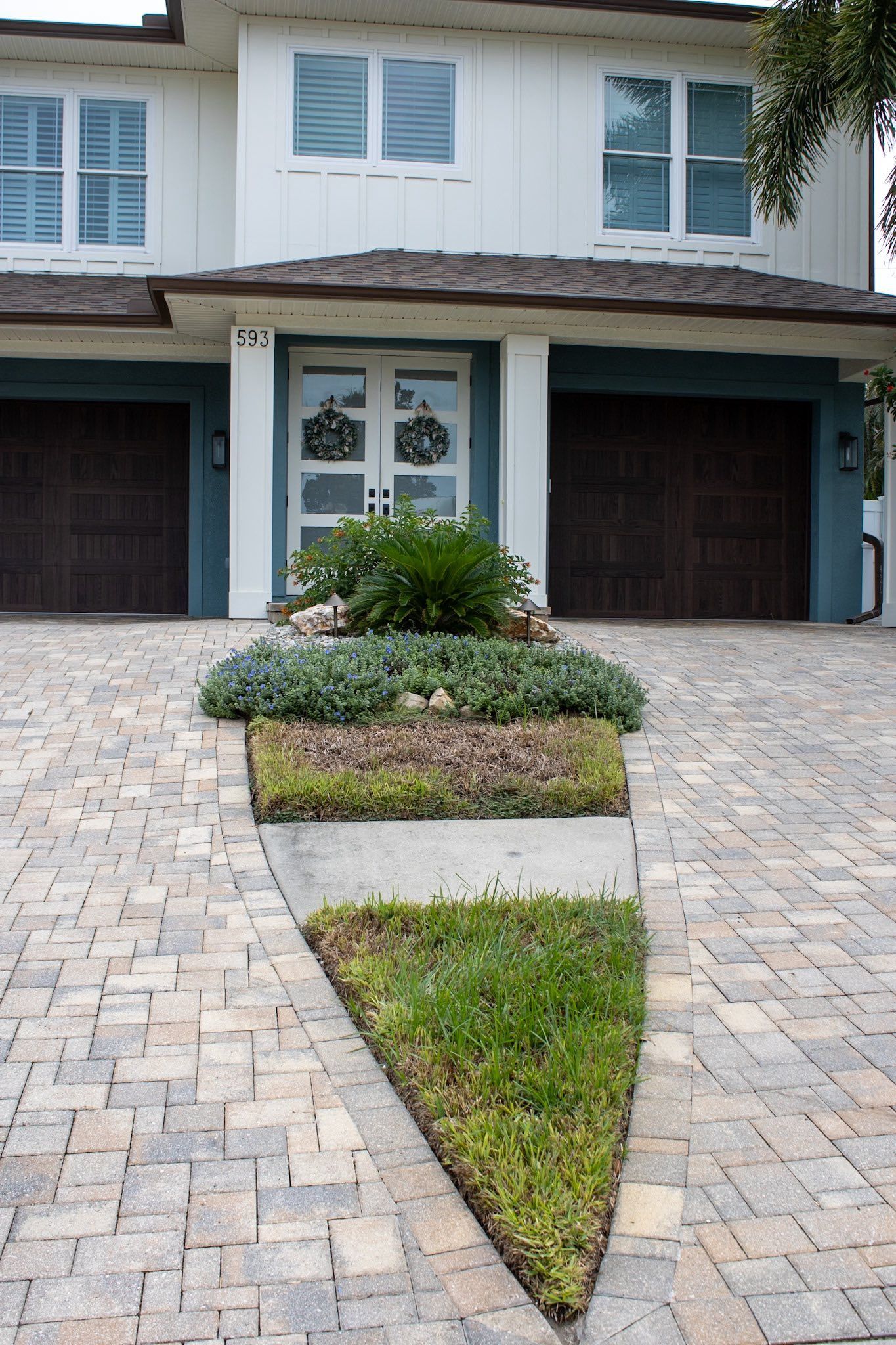 House exterior with driveway and triangular garden bed. Driveway is brick pavers, house is white with blue accents.