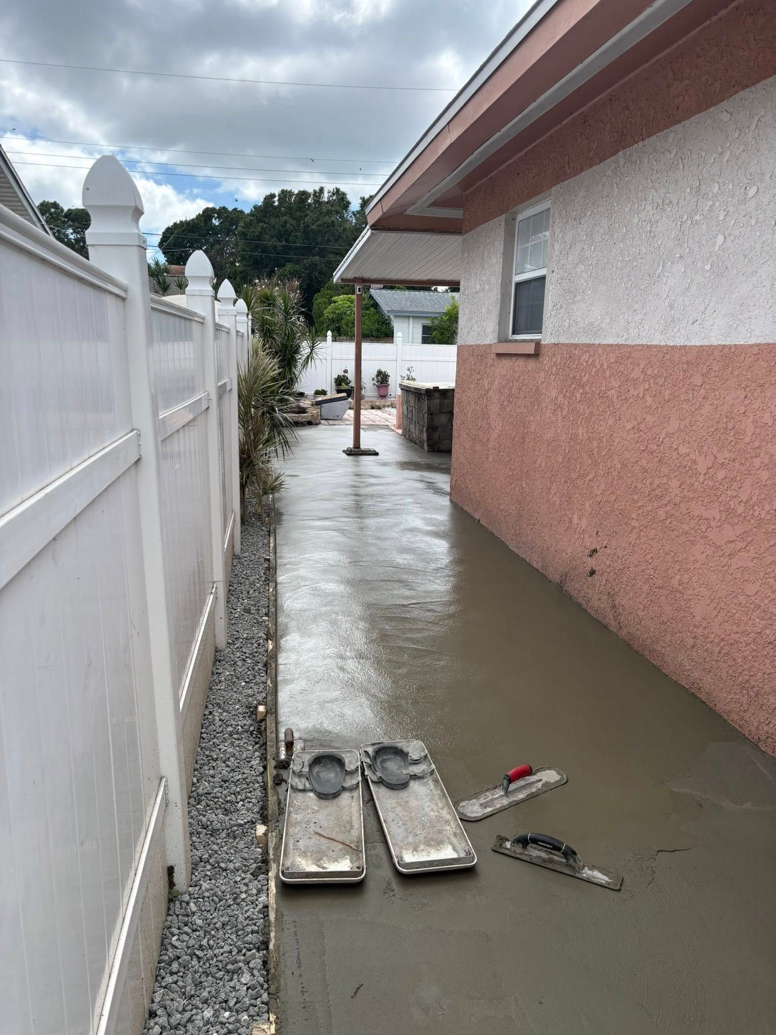 Narrow, concrete alleyway with water, bordered by a white fence and a stucco building. Cementing equipment lies on the ground.