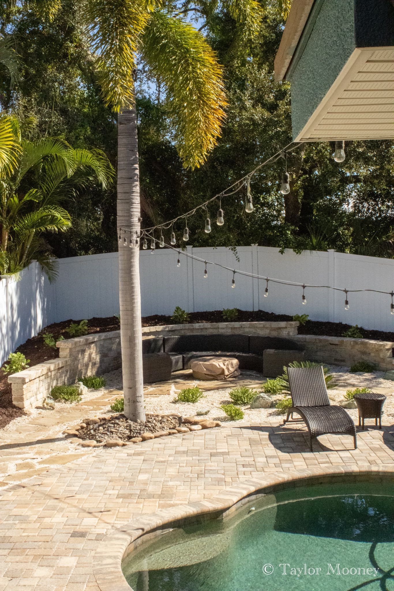 Backyard patio with pool, stone seating area, string lights, and palm tree.