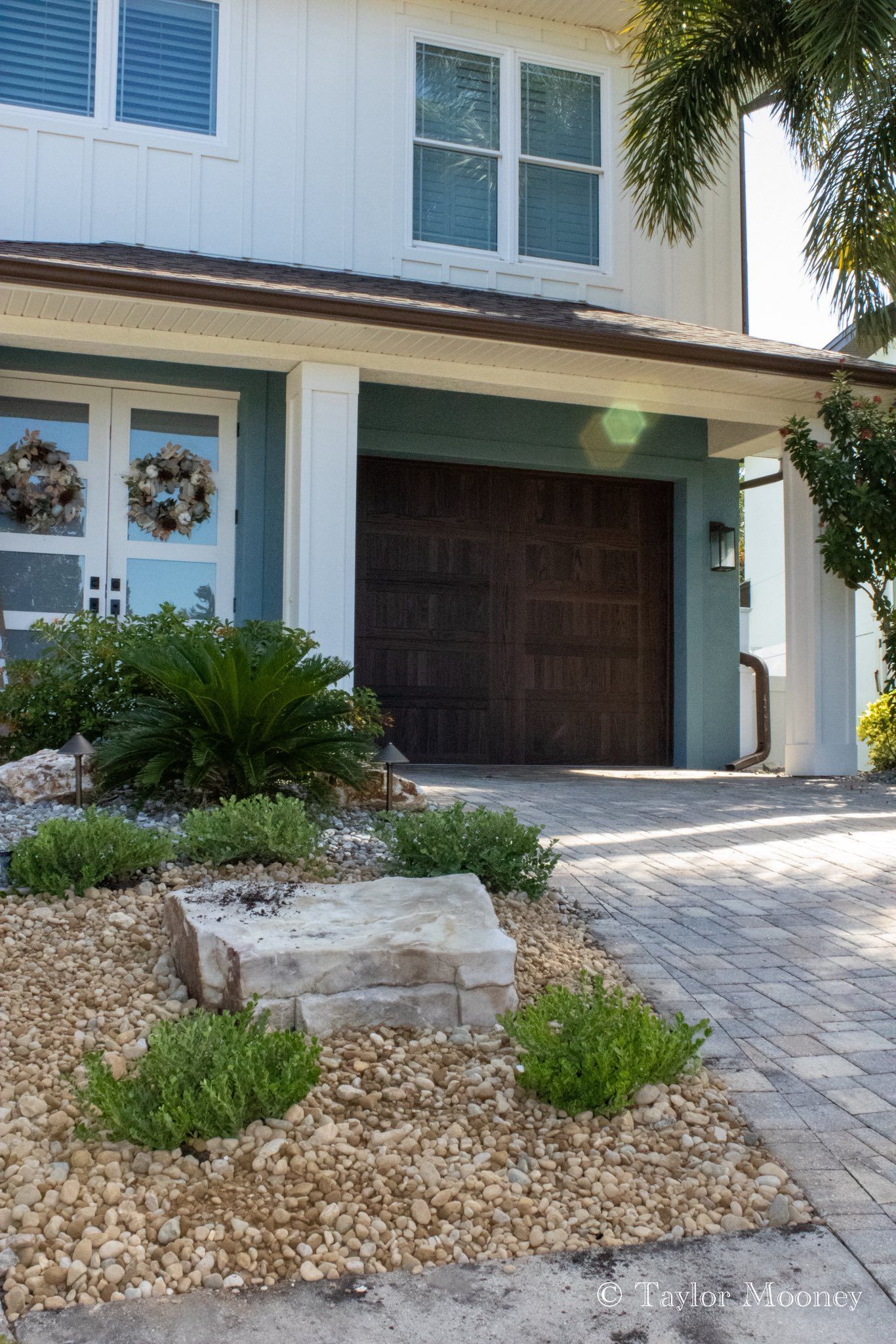 Light blue and white house with brown garage door, rock landscaping, and a paver driveway.