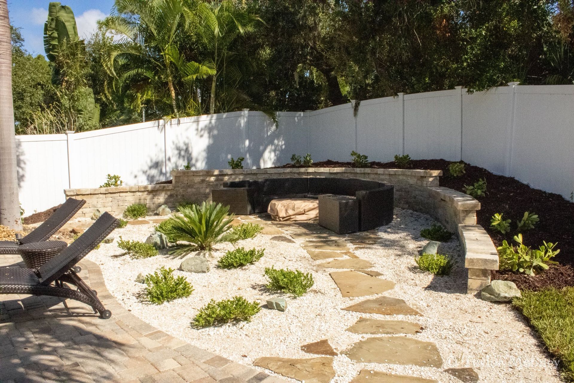 Landscaped backyard with stone fire pit, gravel, stepping stones, and white fence.