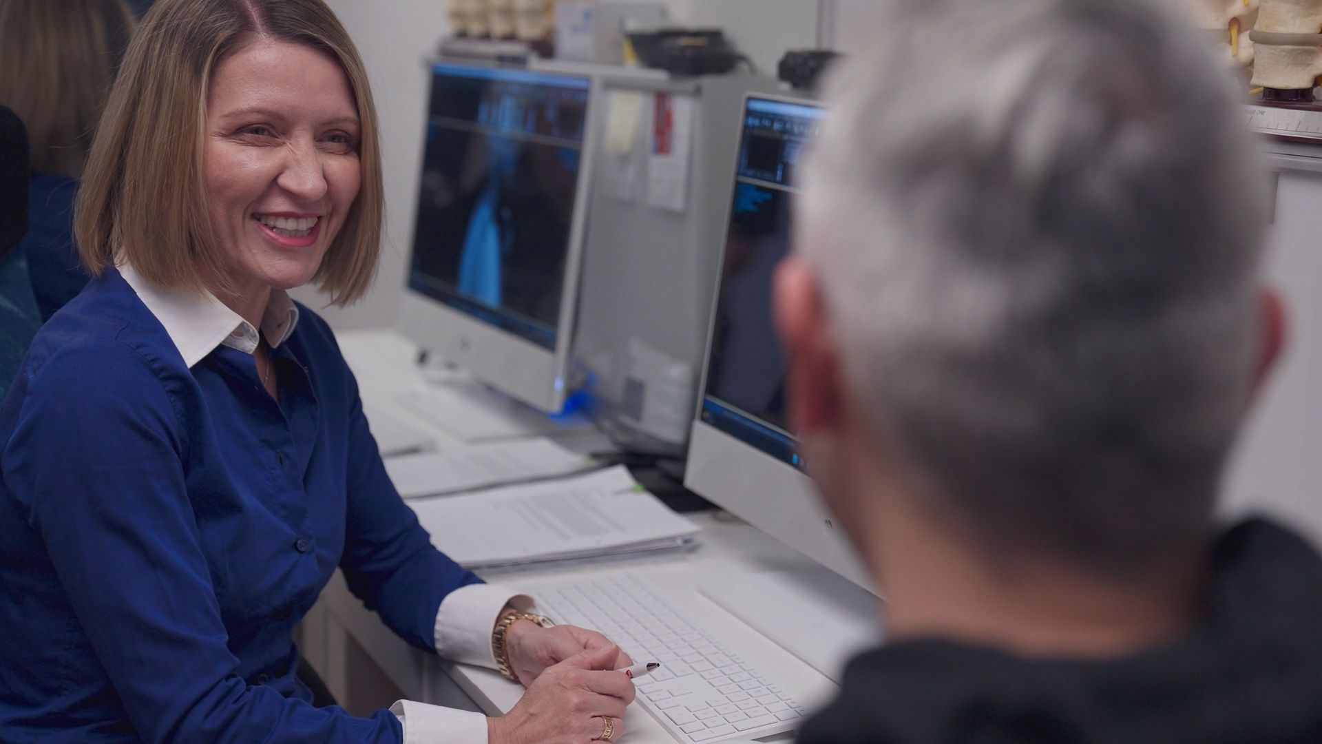 A woman is sitting at a desk talking to a man in front of a computer.