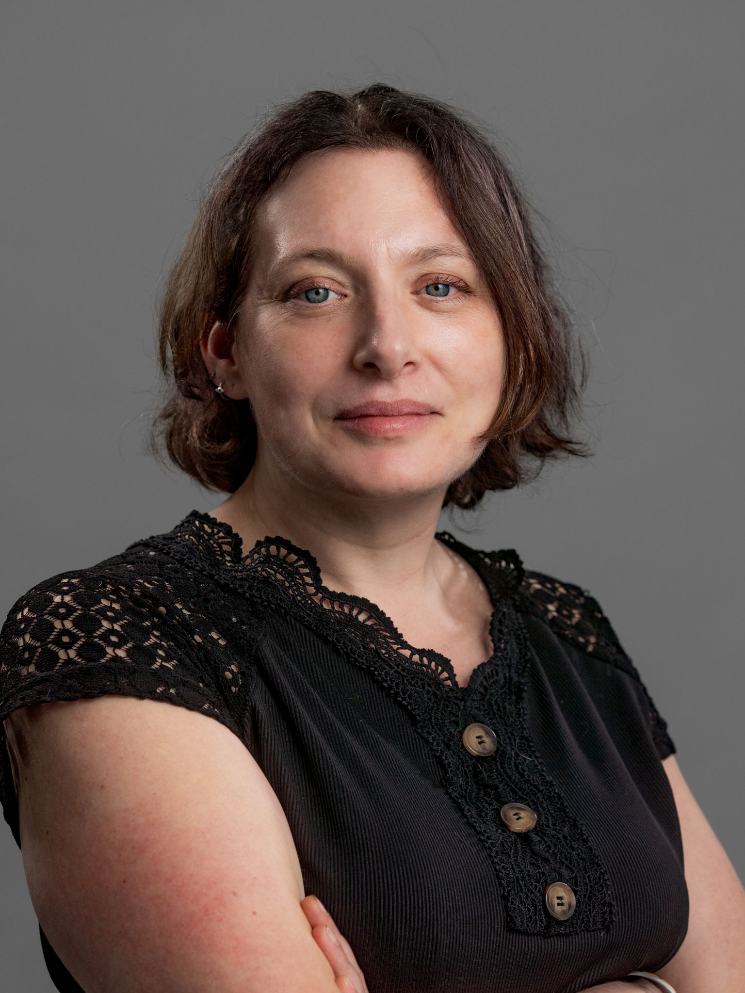 Woman with light skin, short brown hair, wearing a black top with arms crossed, smiling, gray background.