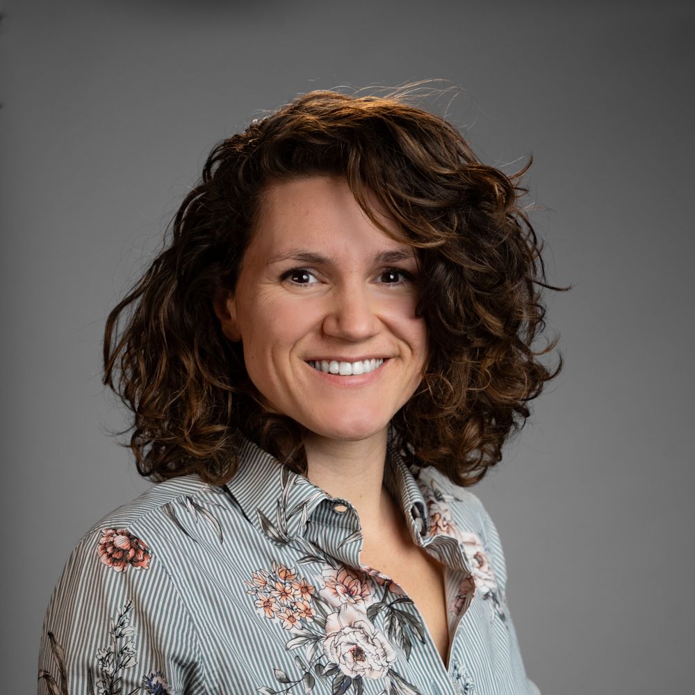 Woman with curly brown hair, wearing a black sweater, smiles gently against a gray background.