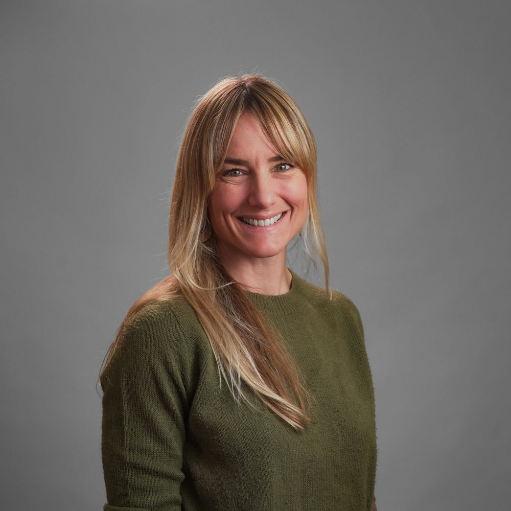 Blonde woman in green sweater smiling at the camera, against a gray background.