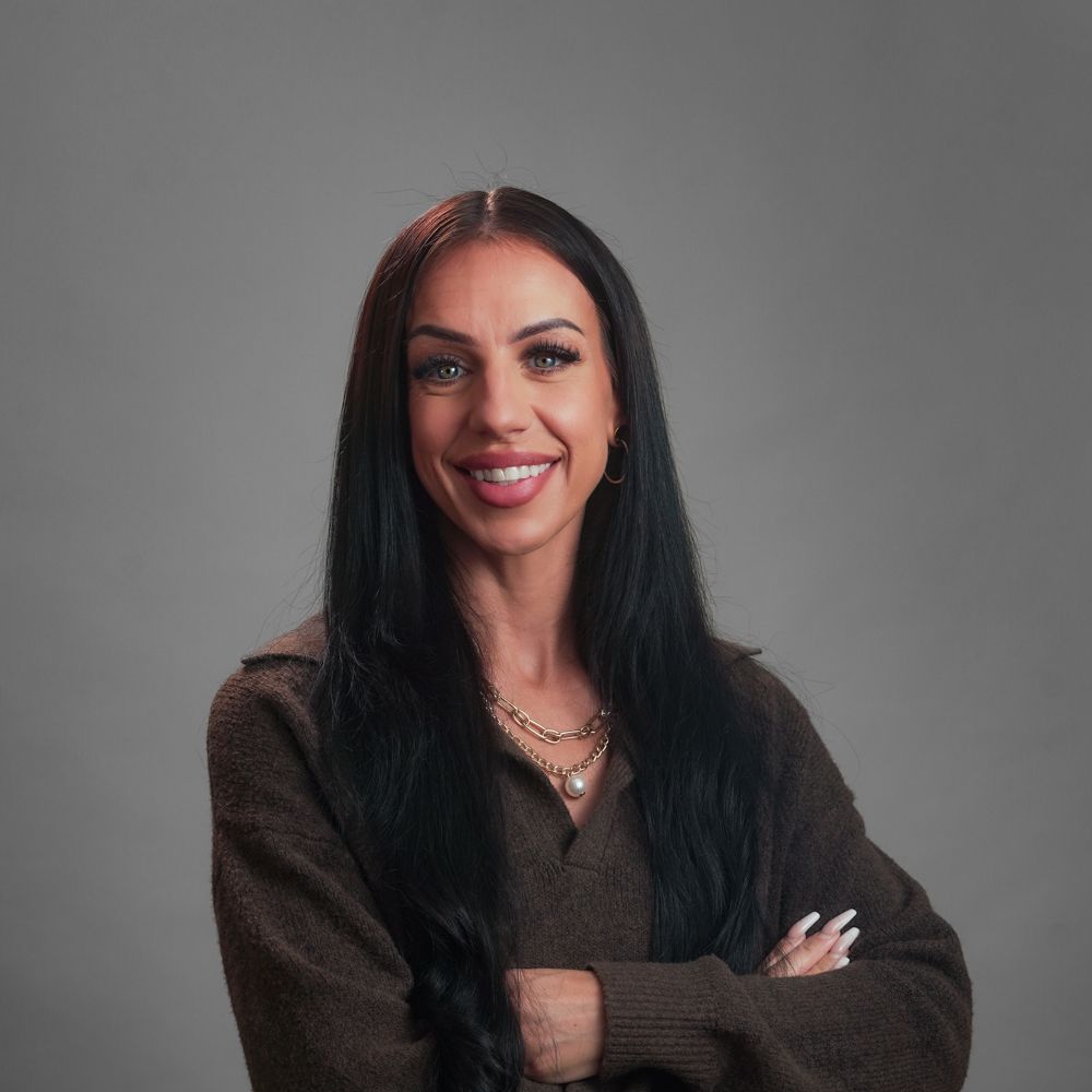 Woman with long black hair, smiling with arms crossed, wearing a brown sweater and gold jewelry.