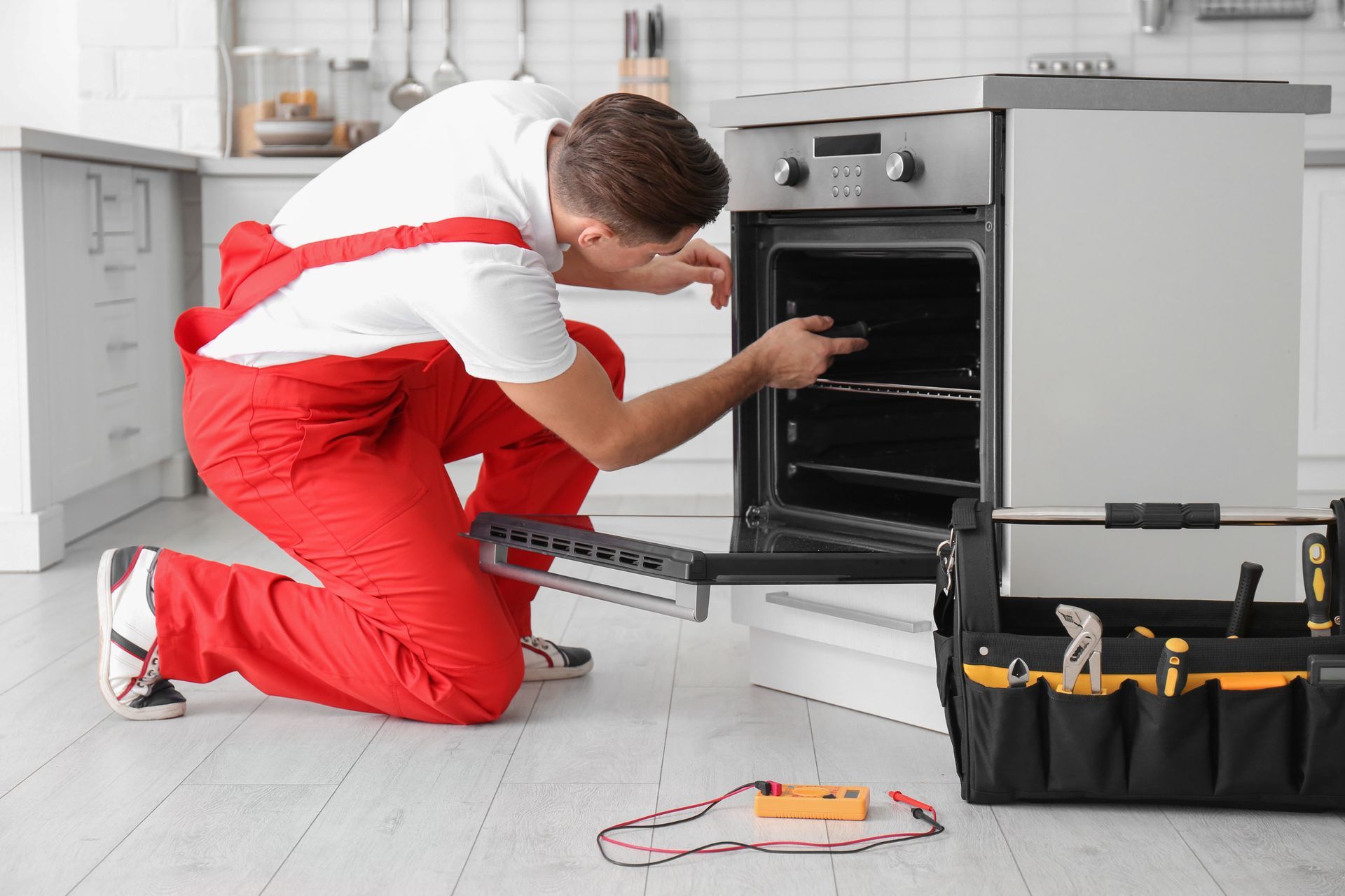 Man in red coveralls repairing an oven in a bright kitchen, using a toolbox and multimeter.