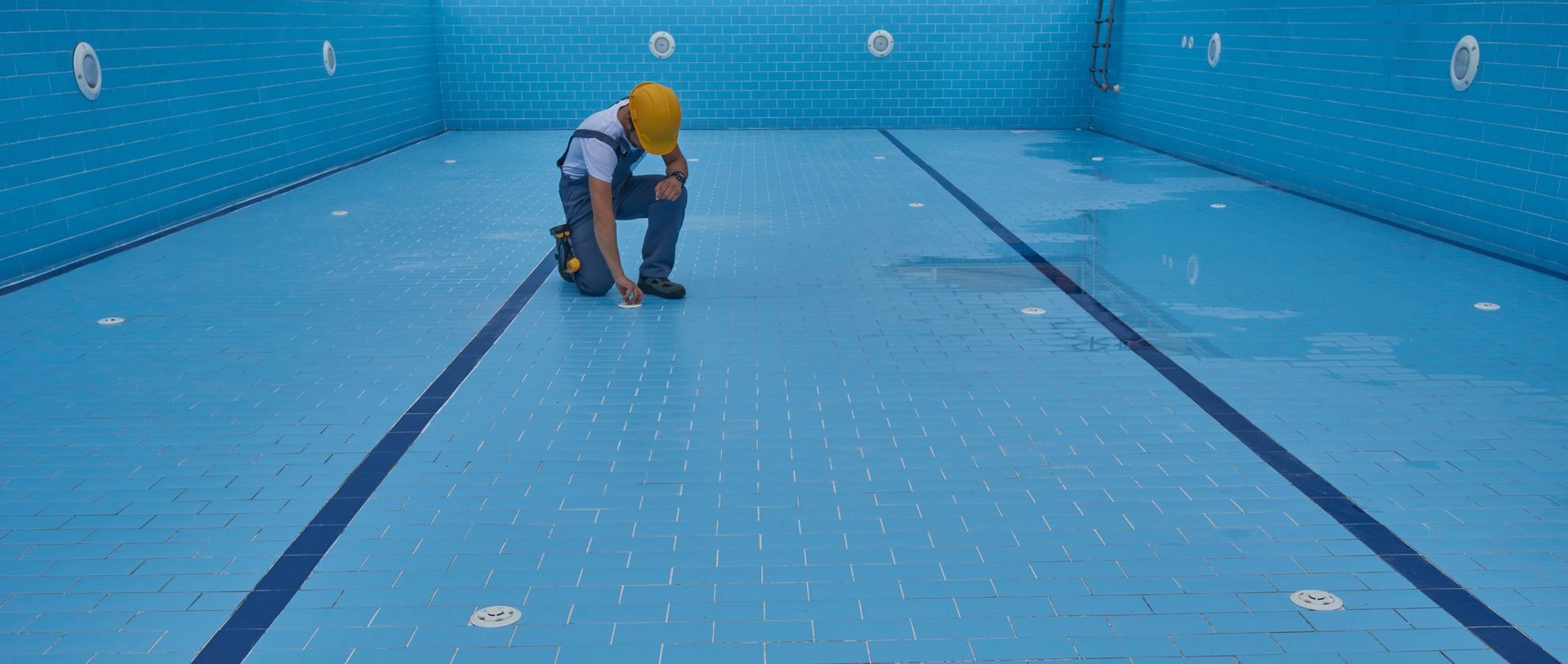 Person wearing a hard hat inspecting the floor of an empty, blue swimming pool.