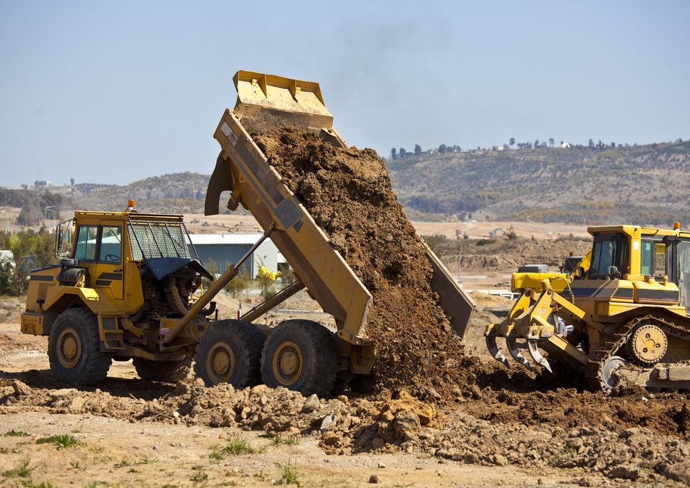 A yellow dump truck in a construction site tilts its bed to deposit a large load of dirt next to a bulldozer.