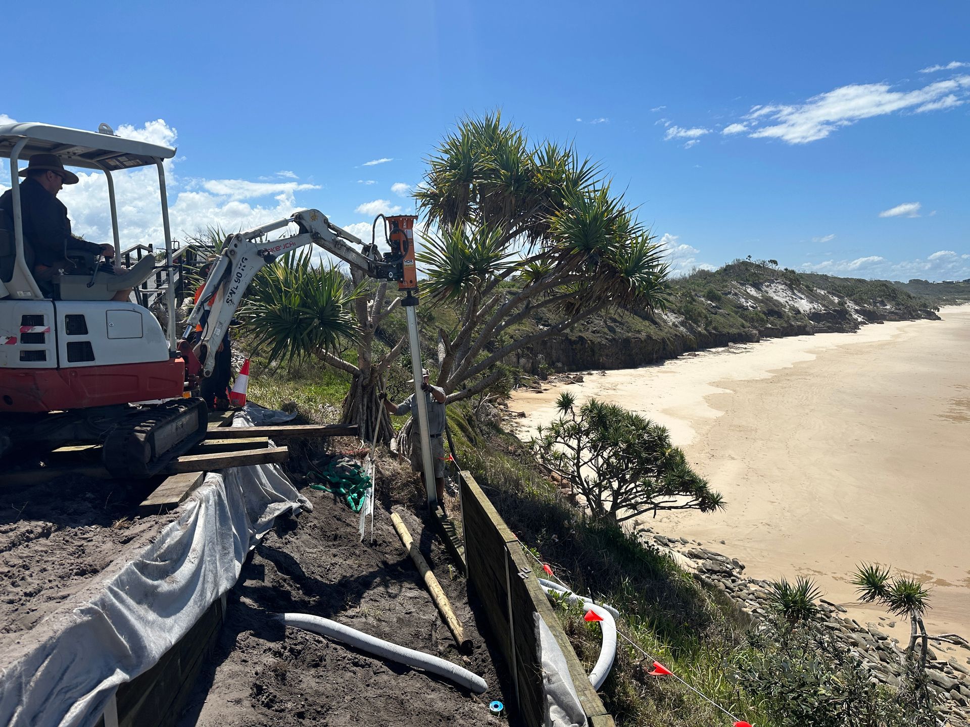 Excavator on a sandy beach beside a fence and palm trees under a clear blue sky