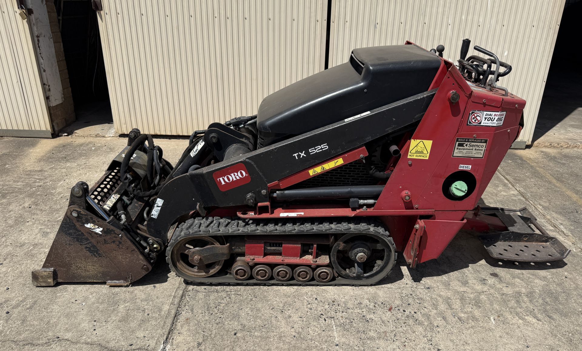 Yellow mini excavator with a black cab on metal tracks, parked beside a corrugated wall.