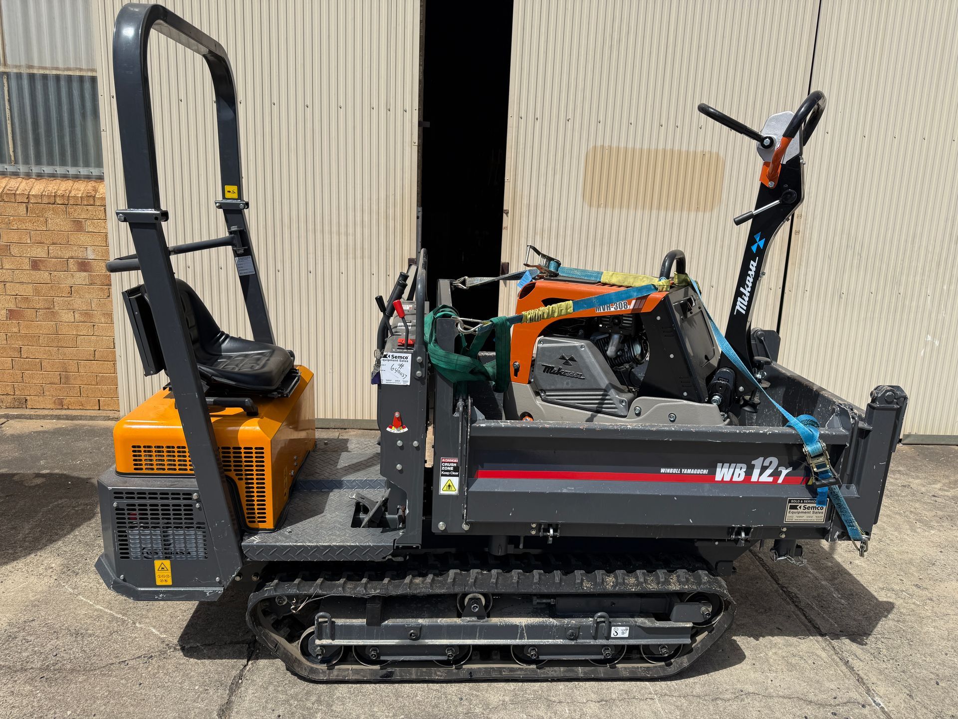 Yellow mini excavator with a black cab on metal tracks, parked beside a corrugated wall.