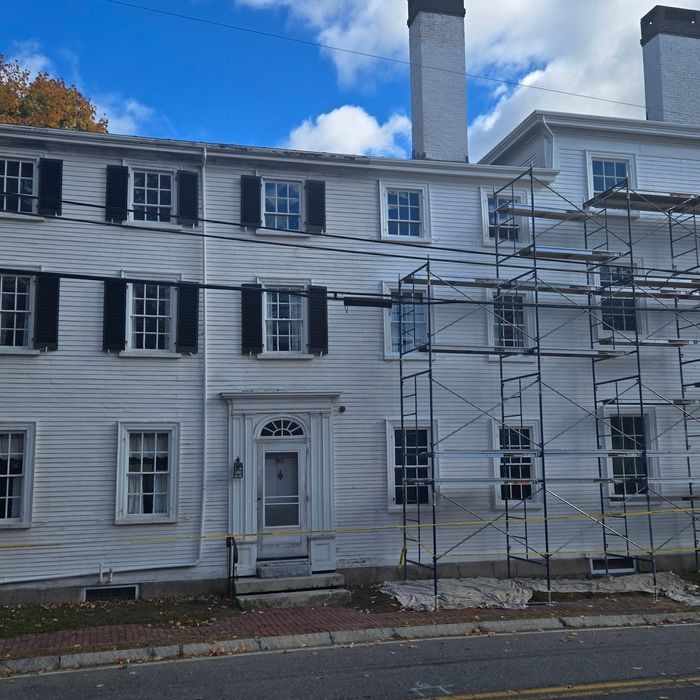 White two-story building with black shutters; scaffolding on the right. Blue sky and autumn foliage visible.