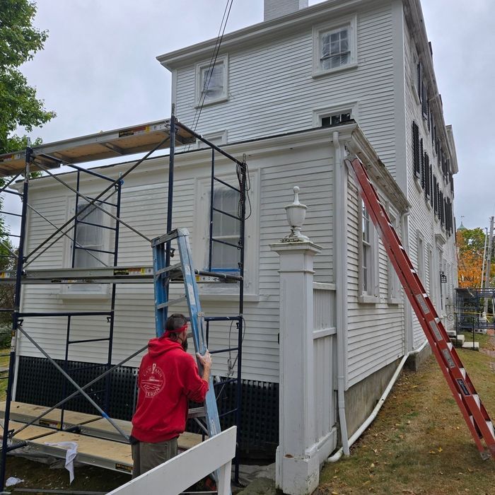 Man in red sweatshirt works on a white house exterior with scaffolding, ladder, and fence. Overcast day.
