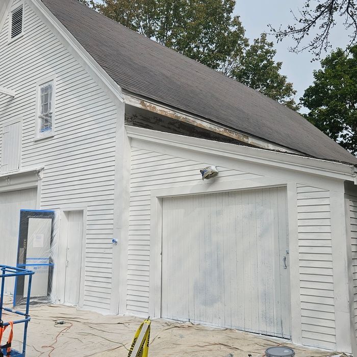 White building being painted; garage door and side entrance are visible, ladder in foreground.