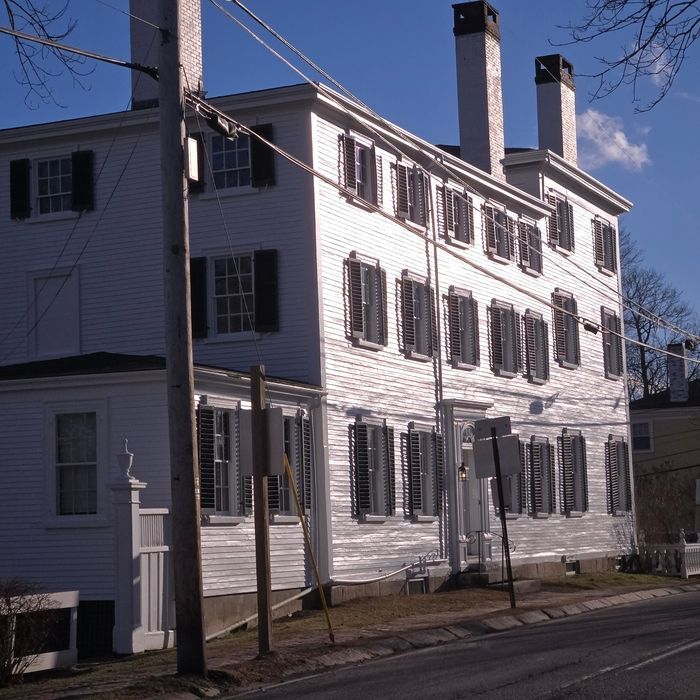 White, three-story building with dark shutters and chimneys, viewed from a street on a sunny day.