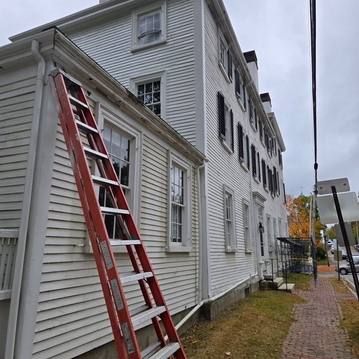 A red ladder leans against a white multi-story building with black shutters on a brick sidewalk.