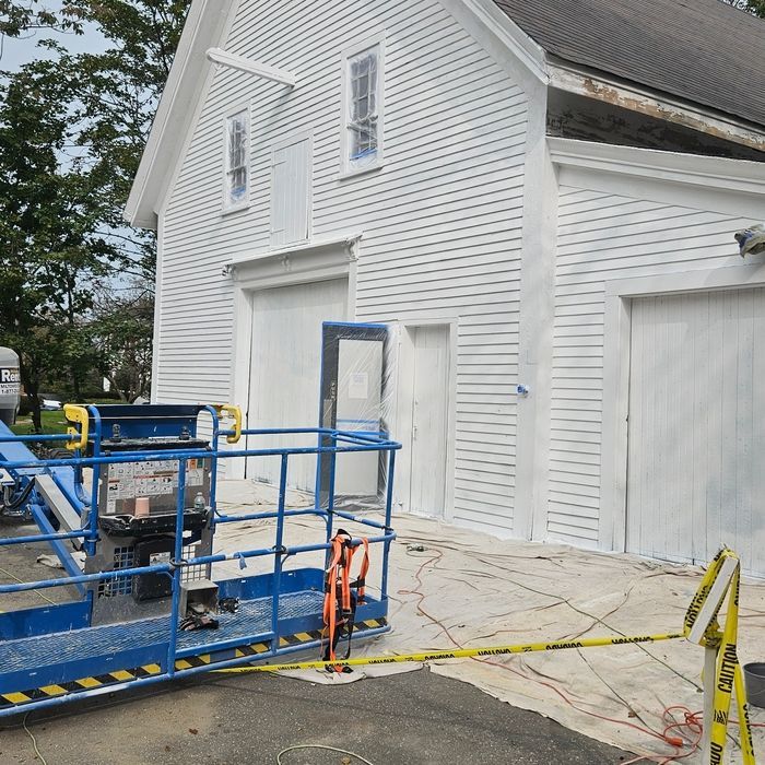 White building with two garage doors and a small door, under renovation. Blue lift, caution tape.