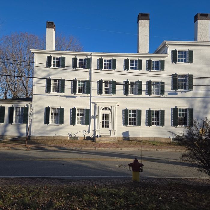 White two-story building with green shutters, black chimneys, and a clear blue sky.