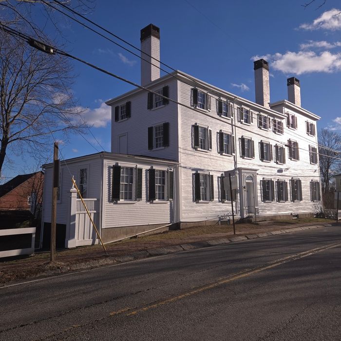 White, three-story building with black shutters and chimneys. Side view, set on a road under a blue sky.