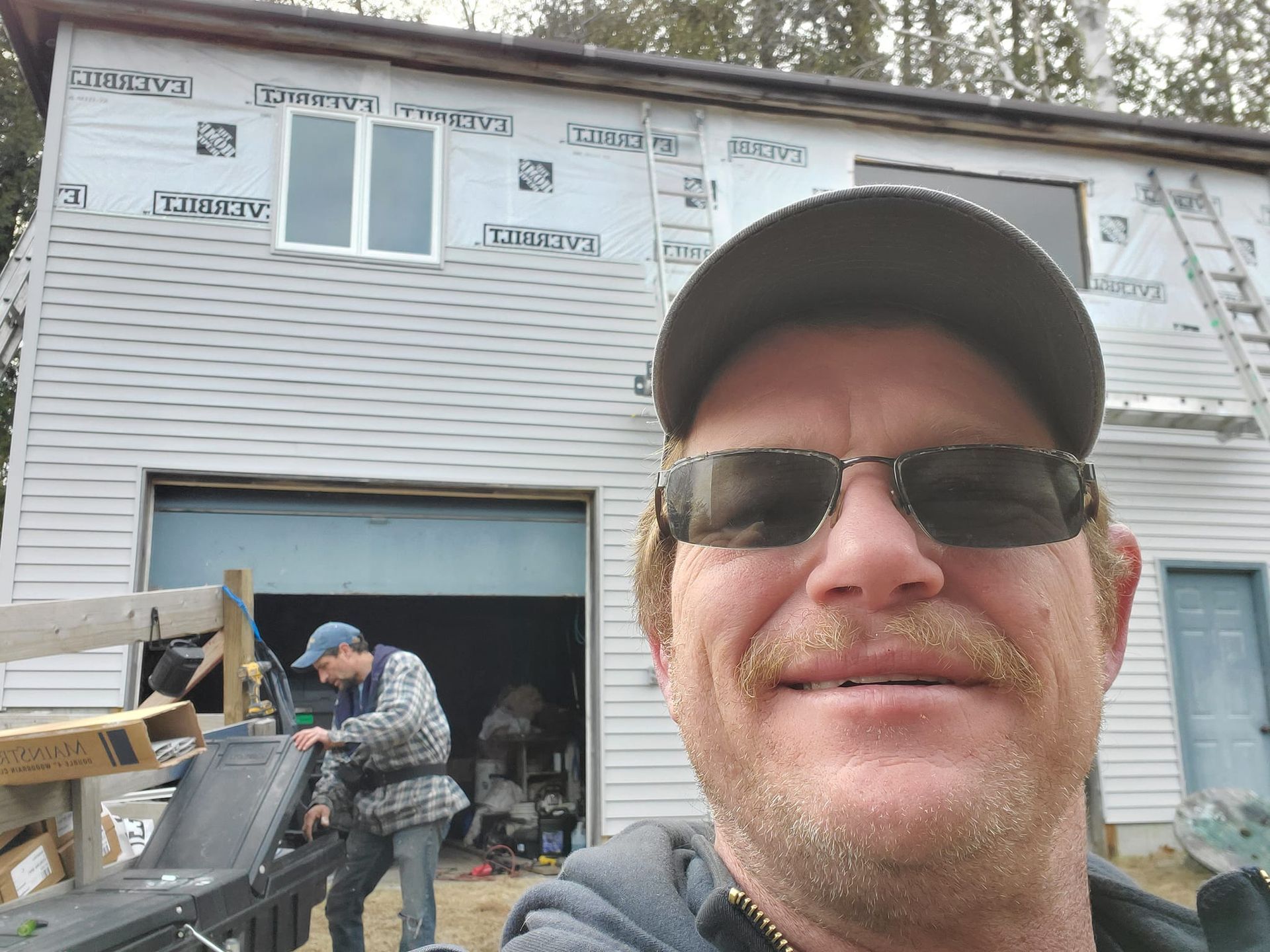 Man in sunglasses selfies in front of a building under construction. Another man works in the garage.