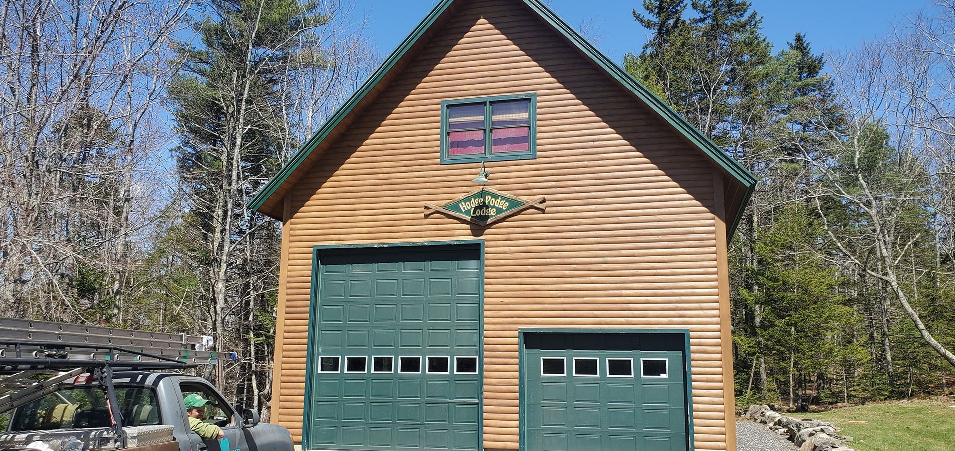 A wooden garage with two green doors, a small window, and a forest backdrop. A vehicle is partially visible.