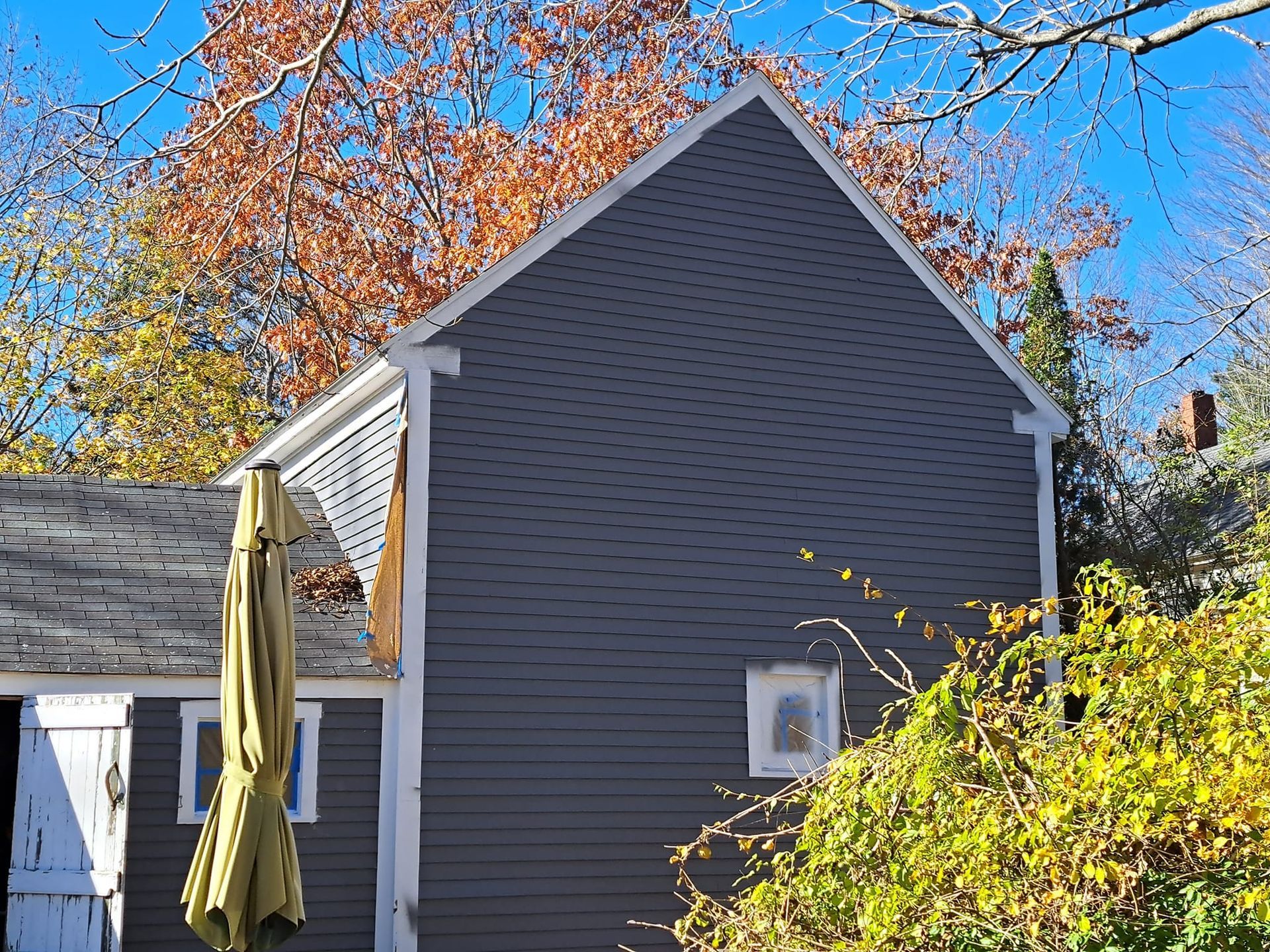Gray house exterior with gabled roof, small window, and white trim against autumn foliage.