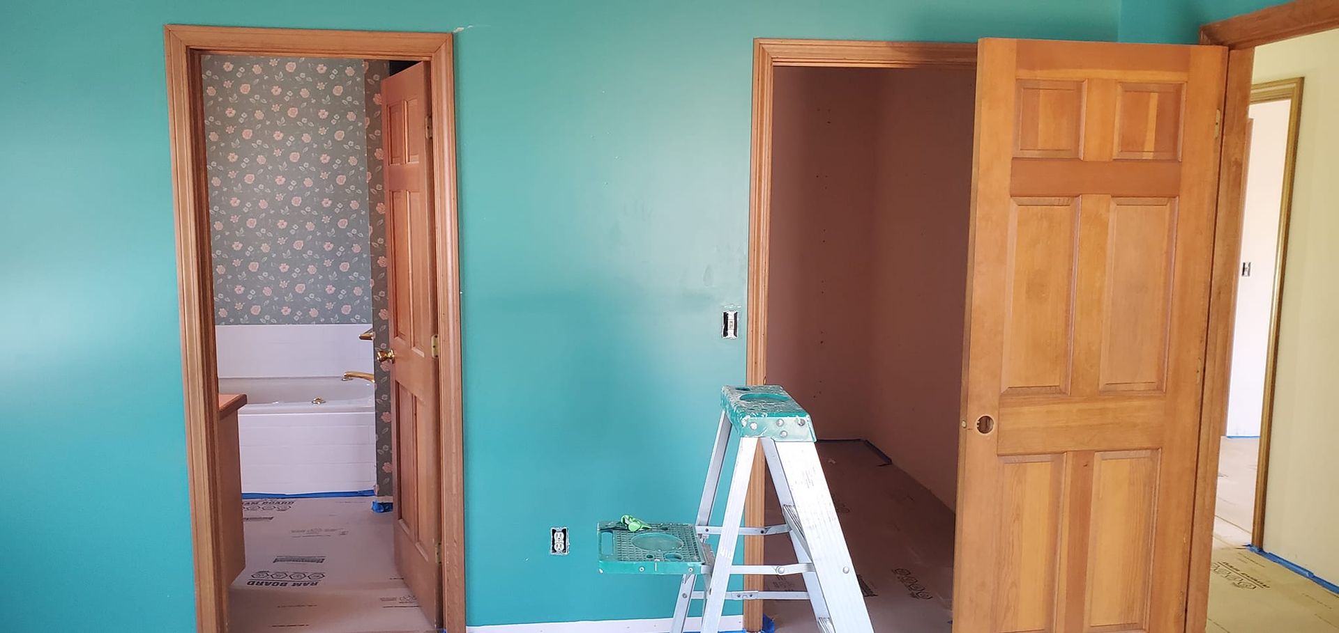 A room with teal walls, wooden door frames, and three open doorways. A stepladder stands in front of one doorway.