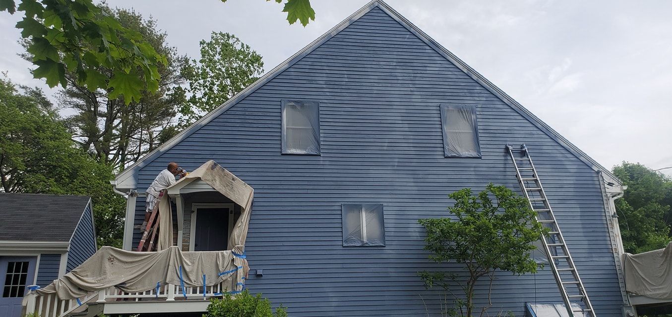 A house is being painted blue. A person on a porch is painting; a ladder leans against the wall.