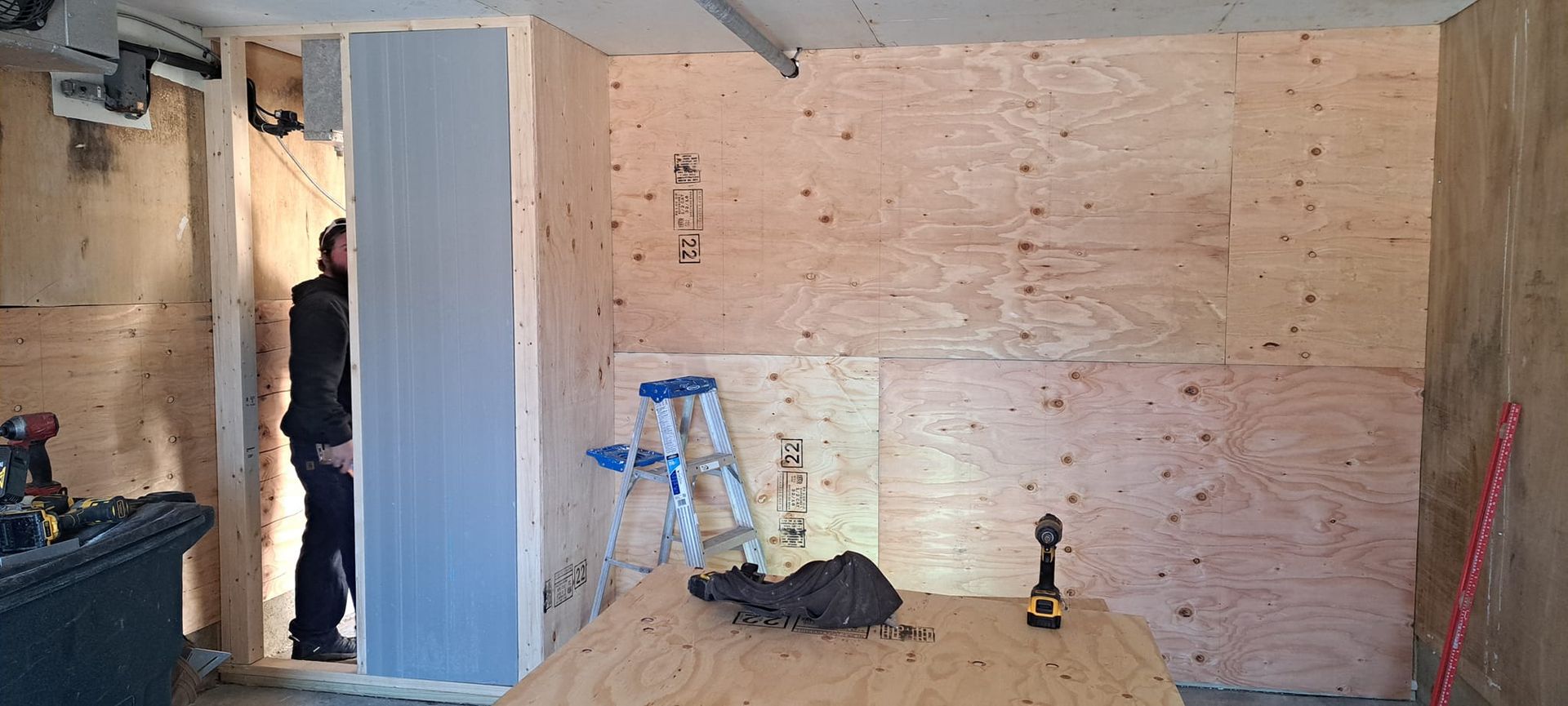 Construction in a room; plywood walls, a person in a doorway, a ladder, and tools are visible.